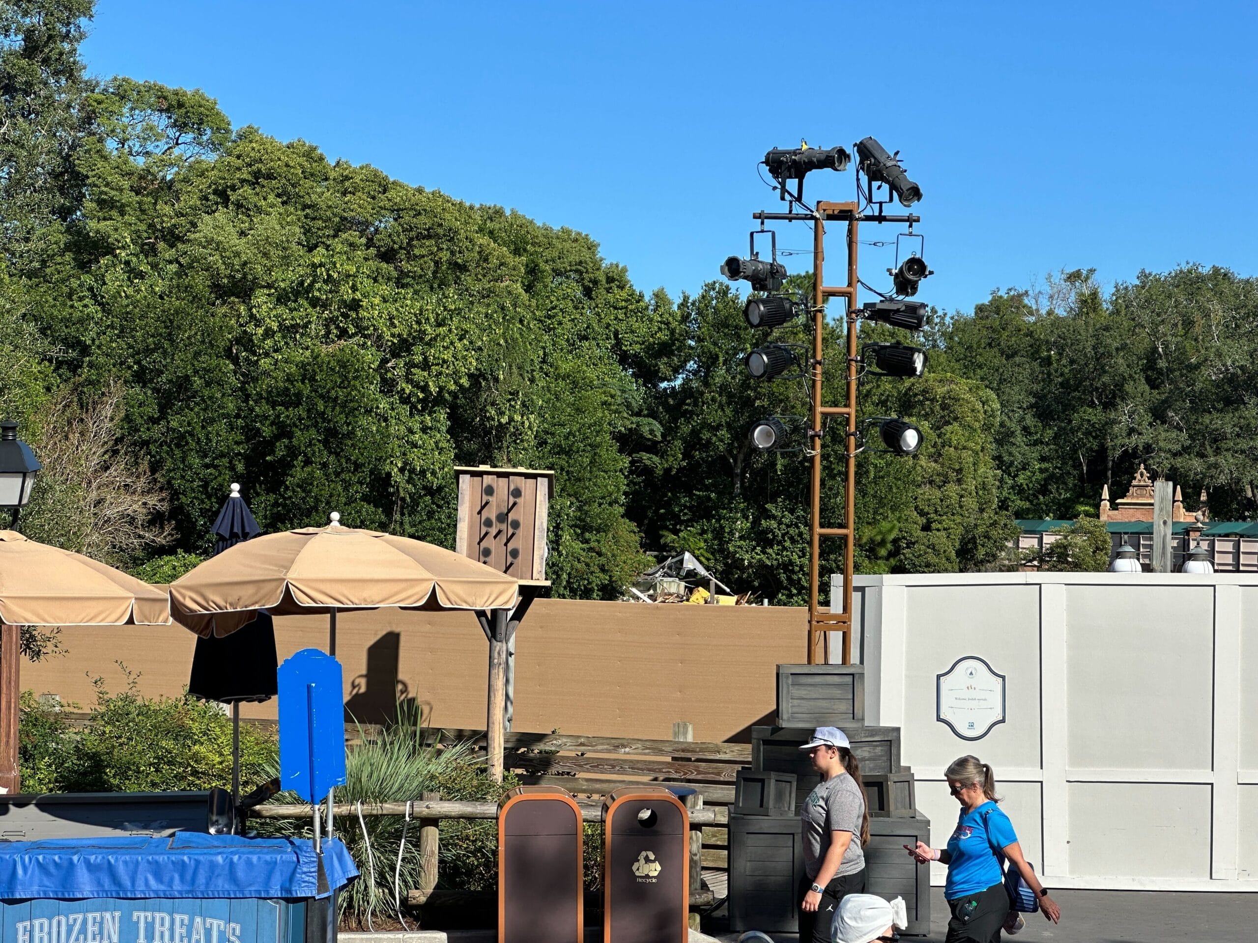 Two guests stroll by construction walls, spotlights, and umbrellas with trees behind—looks like a sunny day in a Disney park.