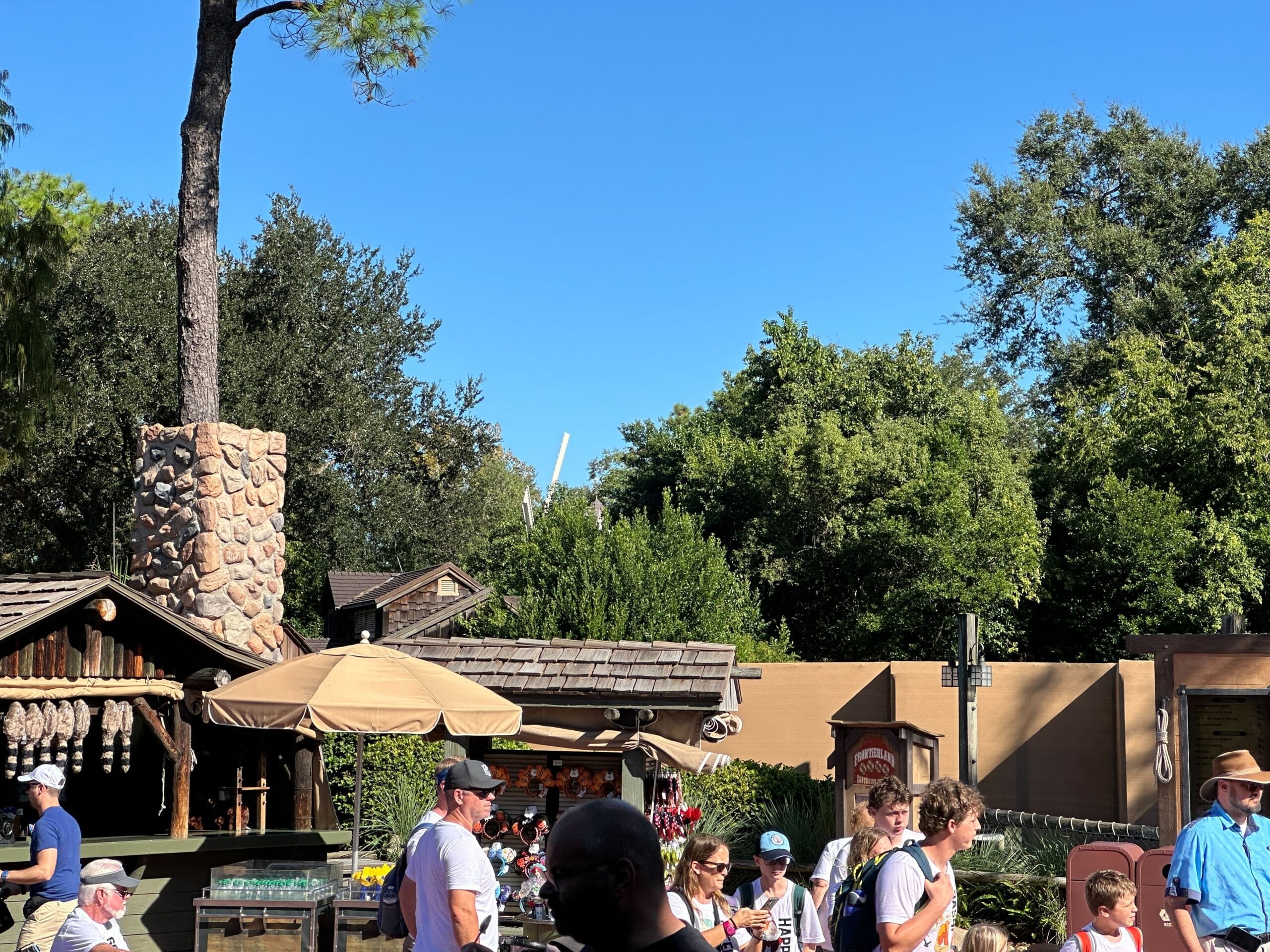 Guests stroll through a lively outdoor market with trees and storybook-style buildings, evoking a Disney park atmosphere, beneath blue skies.