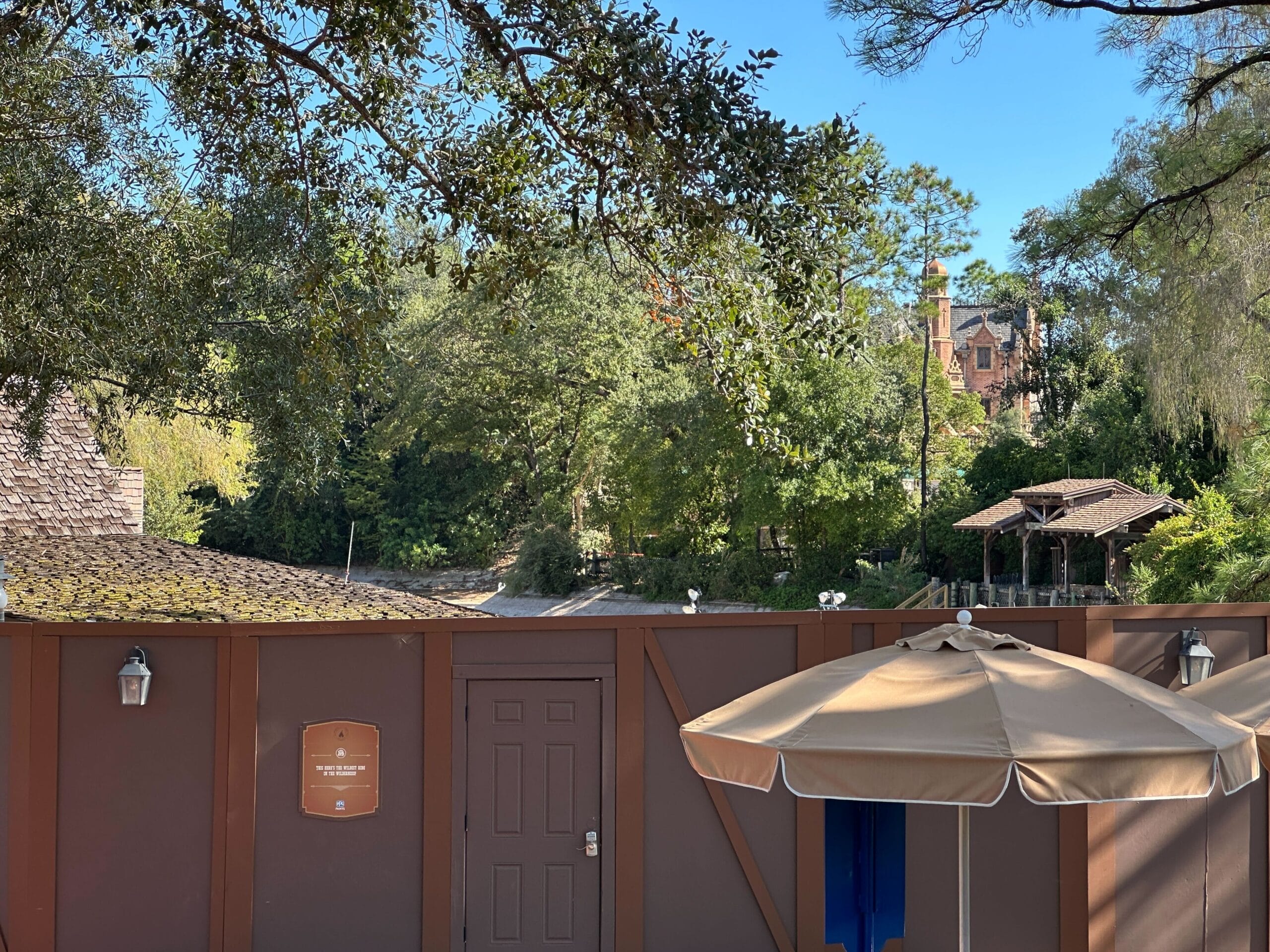 Brown construction wall with a door and beige umbrellas in front; trees and a mansion reminiscent of Disneyland’s Haunted Mansion in the background.