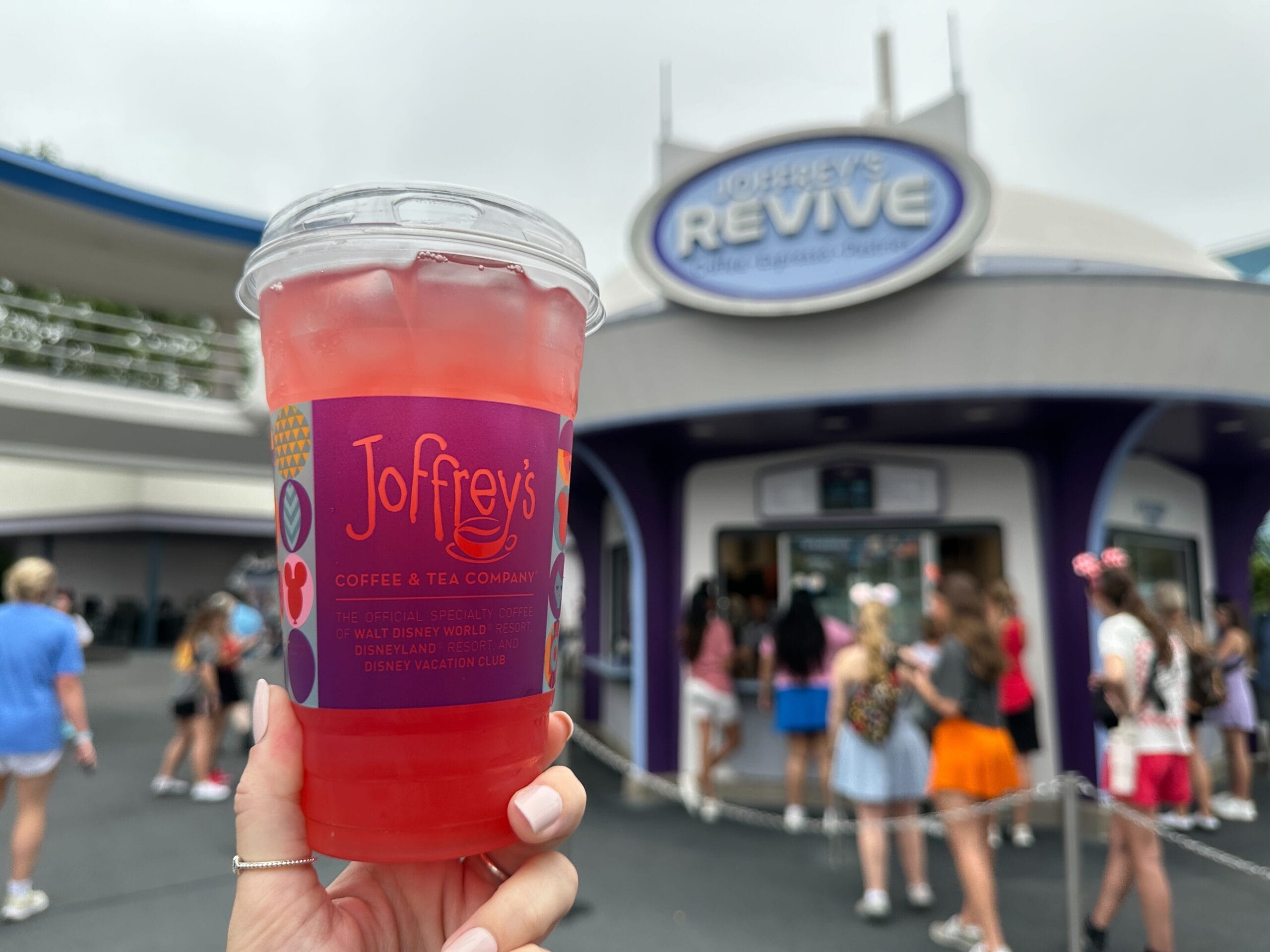 A hand holds a red Joffrey's drink at Magic Kingdom near the Revive café, with TRON Ares Lemonade signs and park guests in view.