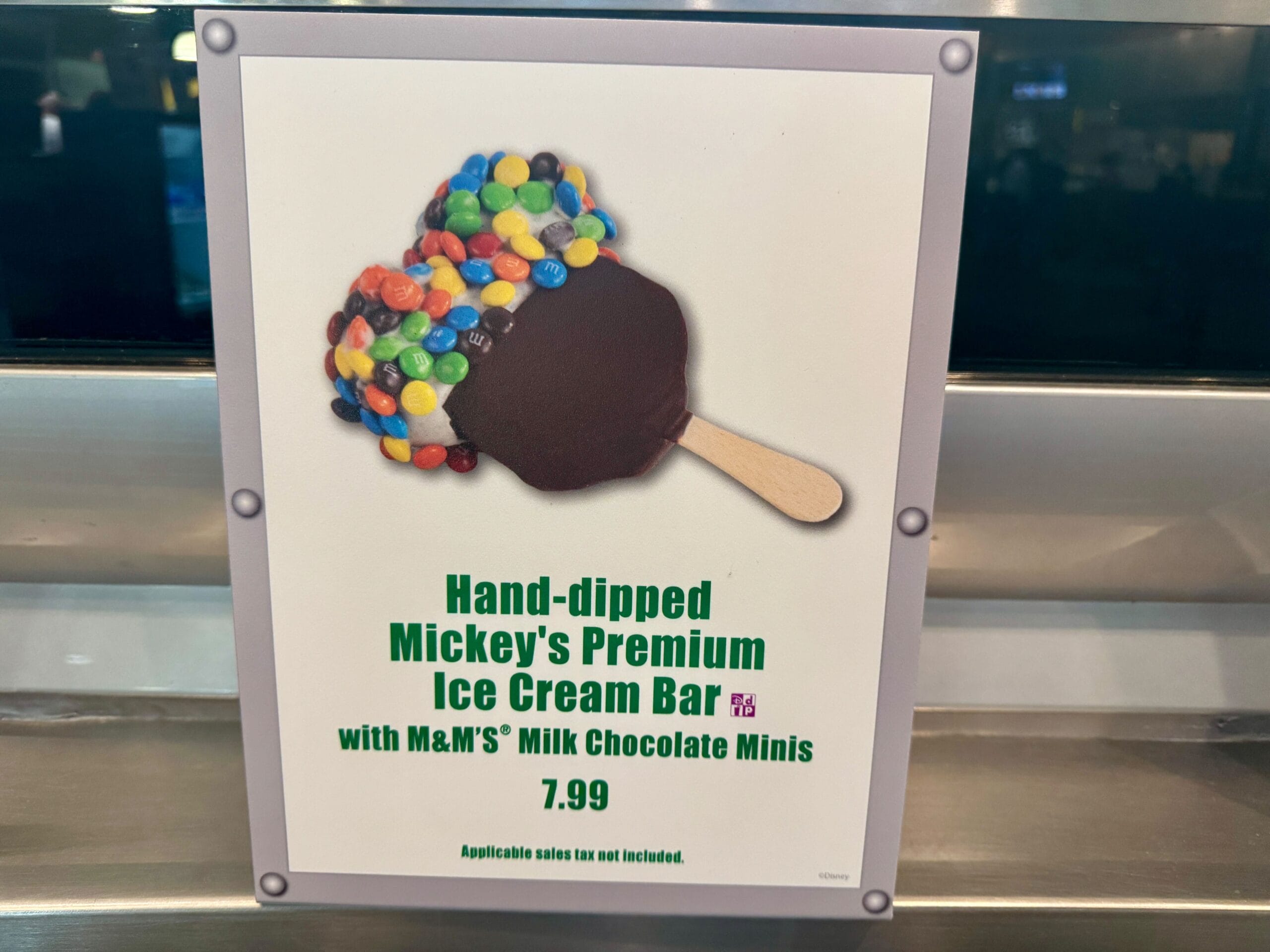 Disney Parks sign featuring a Mickey-shaped ice cream bar with M&Ms, $7.99, labeled "Hand-dipped Mickey’s Premium Ice Cream Bar.