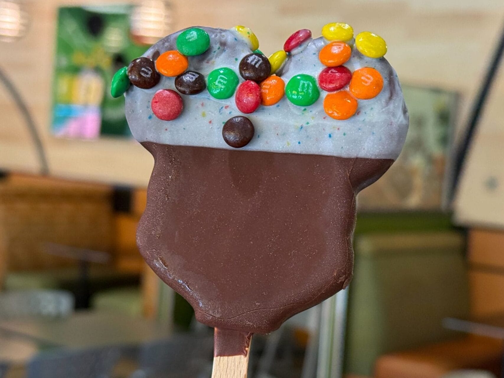 A hand holds a chocolate-dipped ice cream bar topped with colorful mini M&Ms in a modern café at a Disney park.