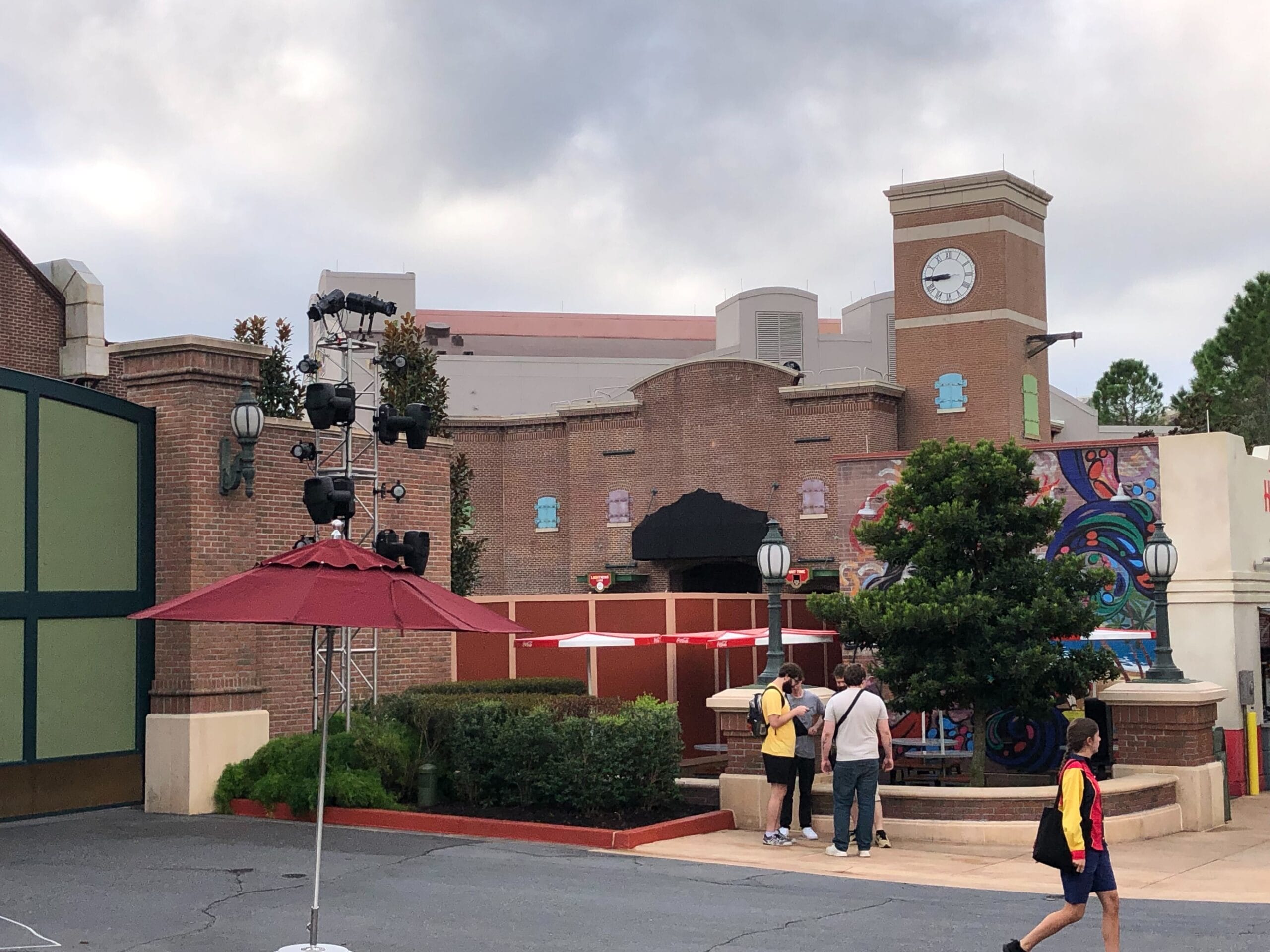 Guests gather near a brick building with a clock tower, greenery, and red umbrella—reminiscent of Main Street in Disney Parks.