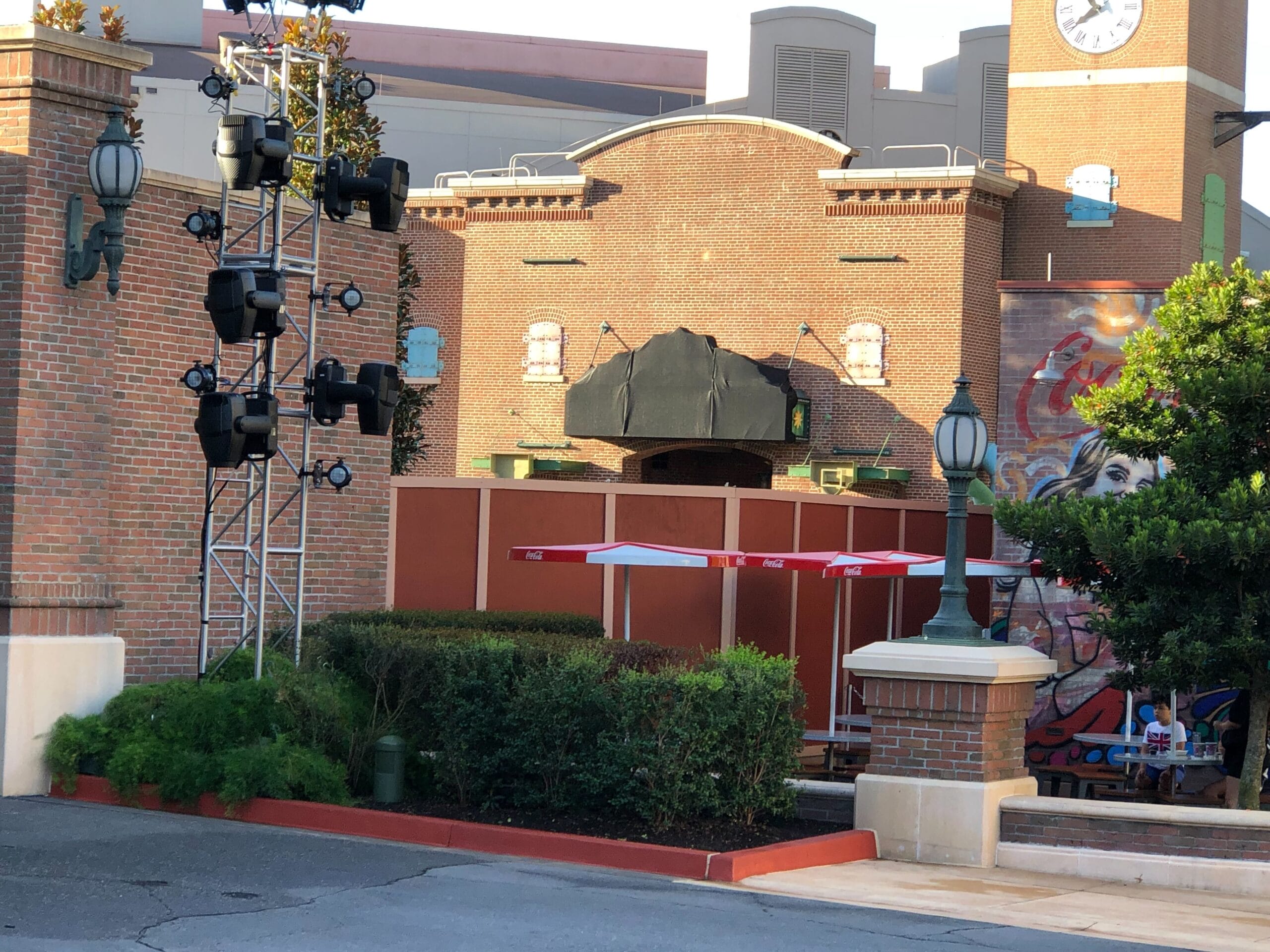 Outdoor patio with red umbrellas near a brick building and mural, partially hidden by construction barriers—does not resemble Disney castles.