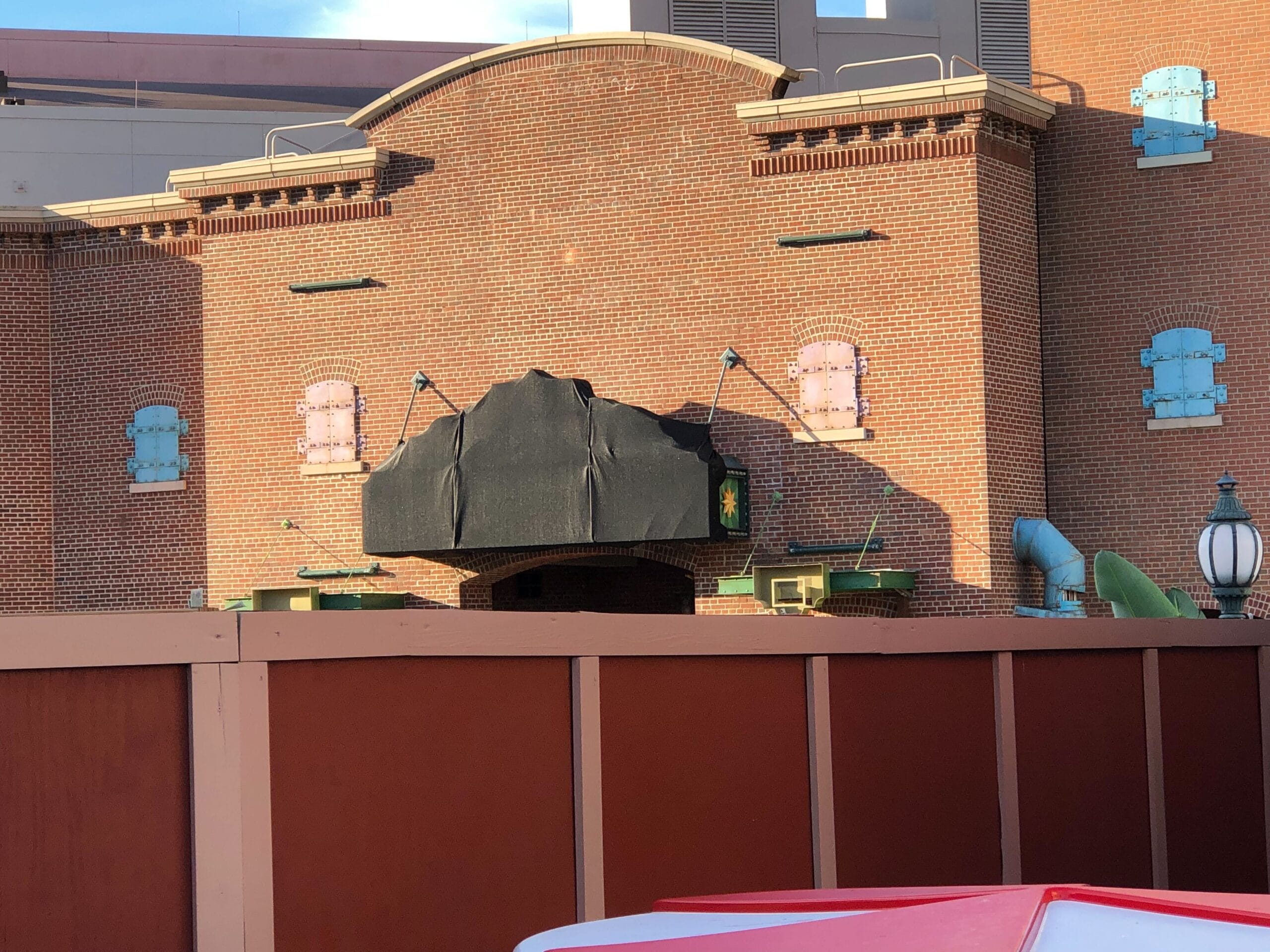 A whimsical brick building with a covered marquee and bright shutters, partially hidden by a red construction fence, Disney park style.