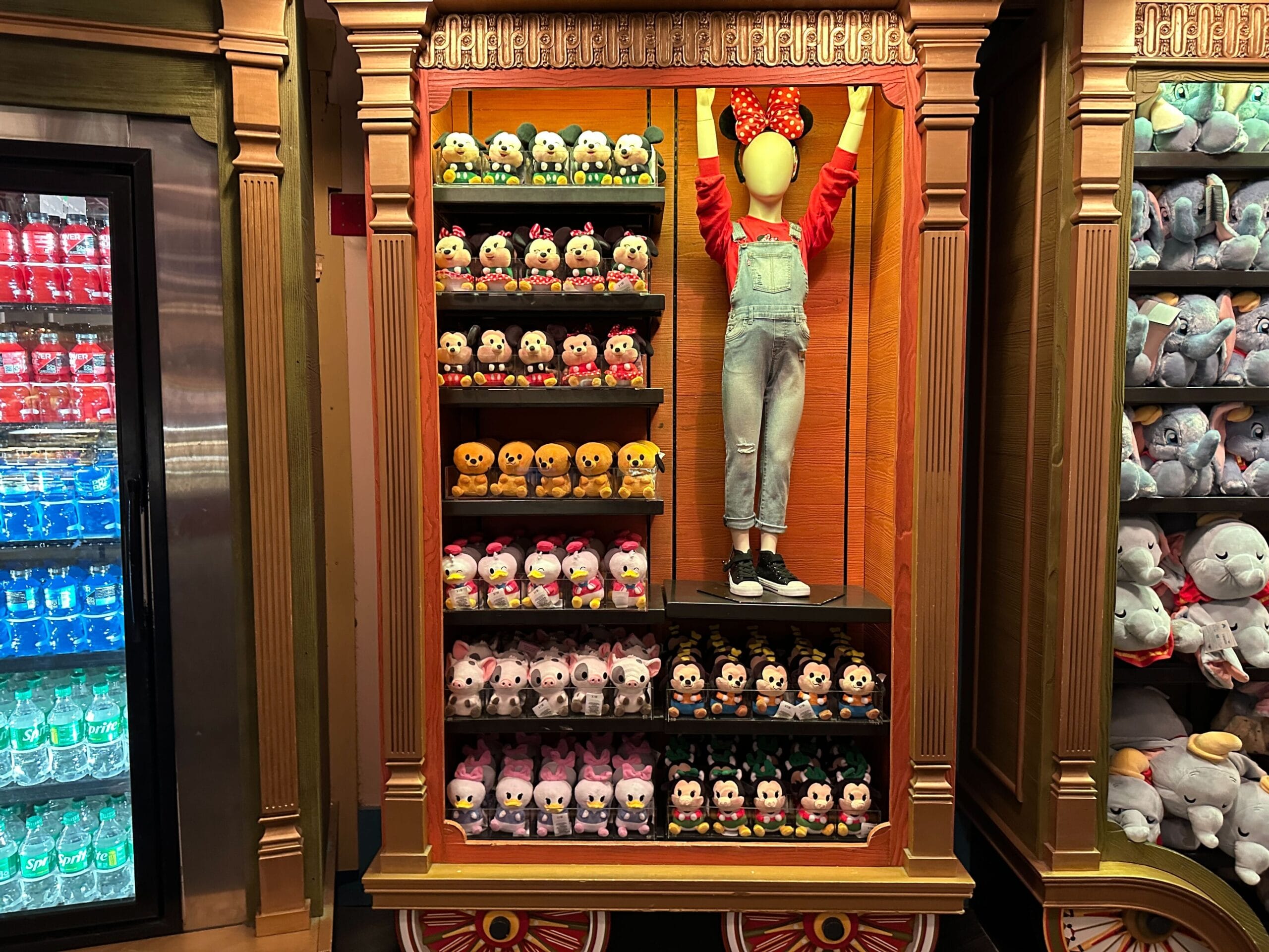 Plush Disney toys and a mannequin in Minnie Mouse ears line store shelves by a fridge with drinks, evoking a Disneyland gift shop.