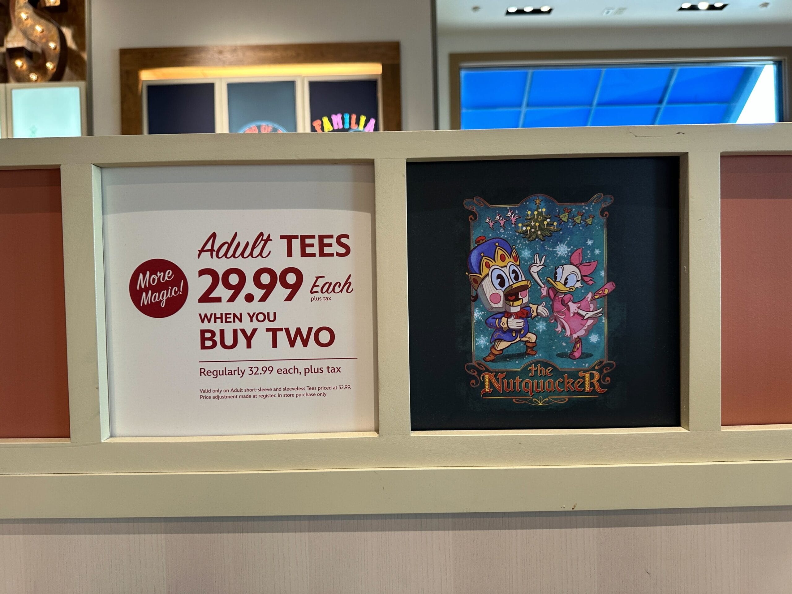 Sign at a Disney park selling adult tees for $29.99 each with purchase of two, beside a "The Nutcracker" Disney movie poster.