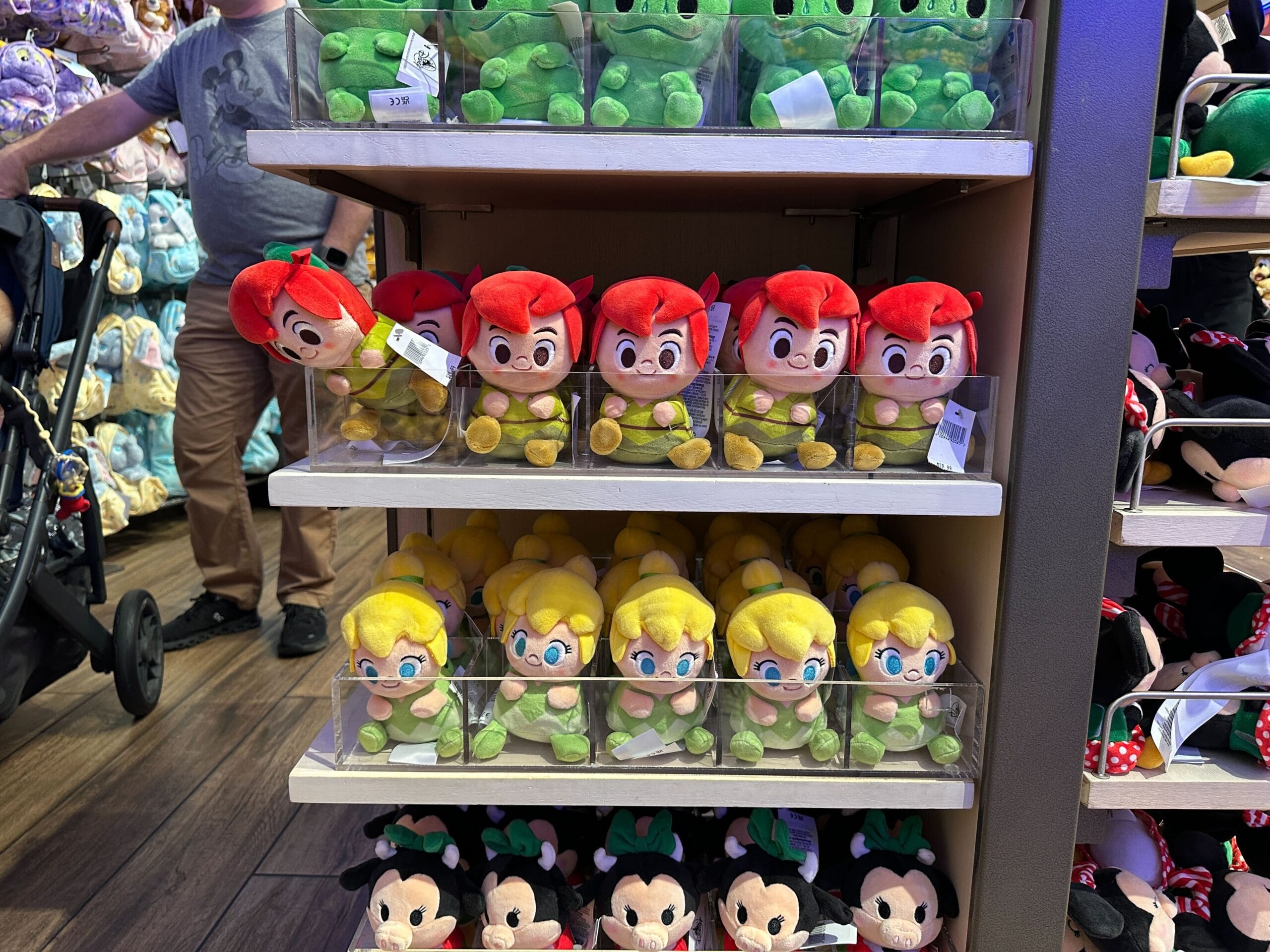 Plush Peter Pan character dolls on shop shelves at a Disney Park, with shoppers and strollers, evoke park merchandise magic.