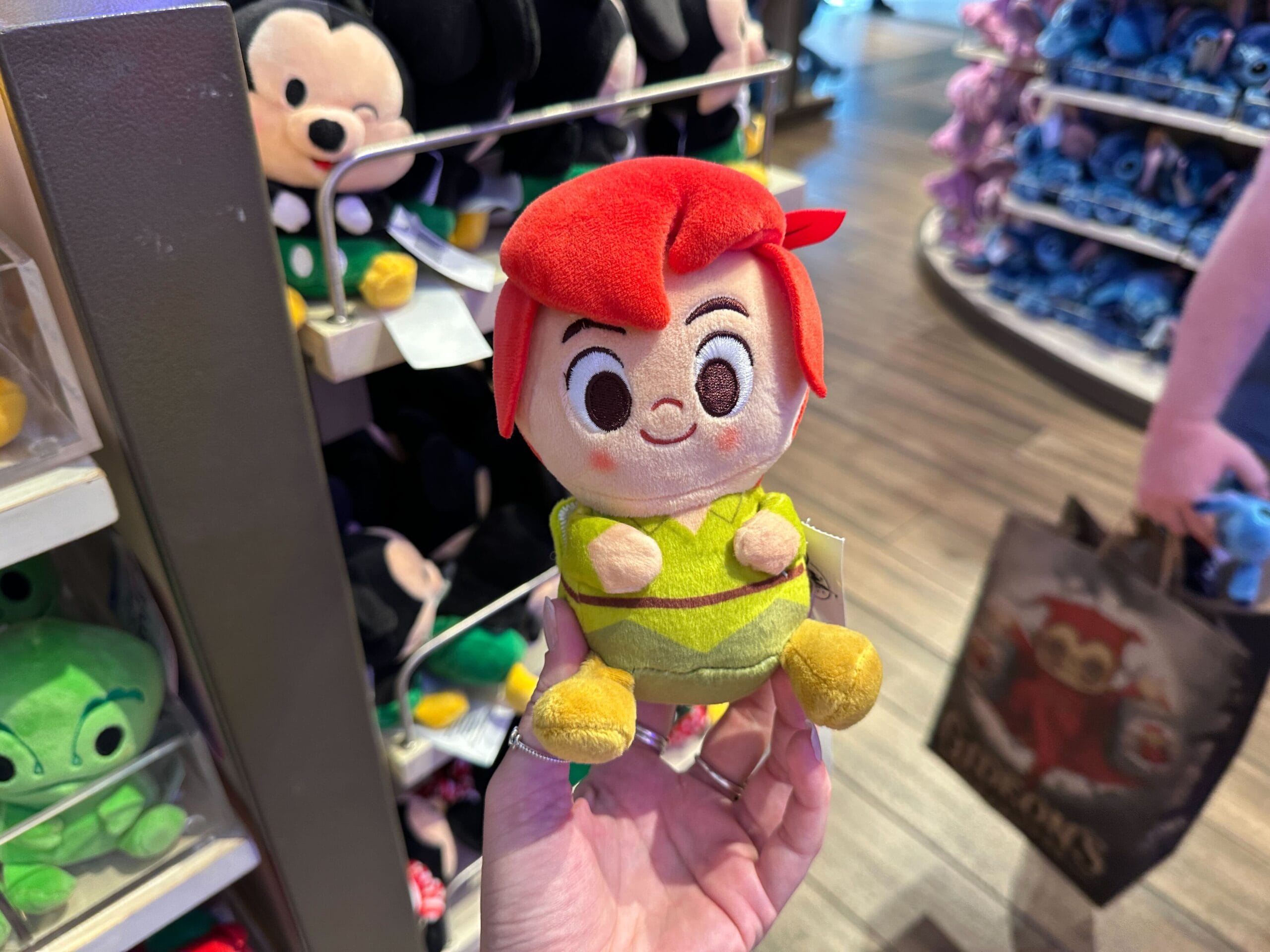 A hand holds a small Peter Pan plush toy in a Disney park store, with other plushies, like ones found at Main Street shops, behind.