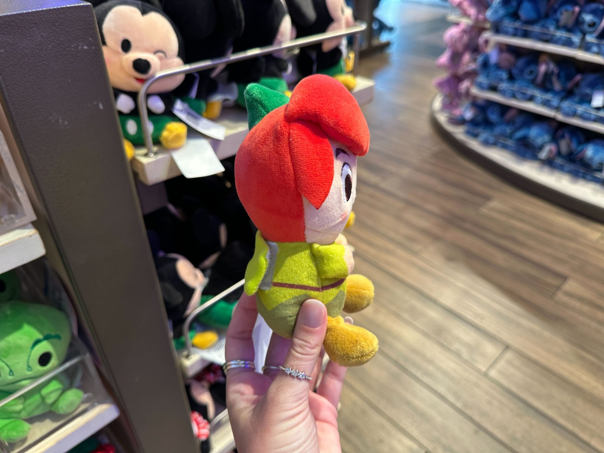 A hand displays a plush toy in a red flower outfit, surrounded by more plushes on shop shelves at a Disney park merchandise store.
