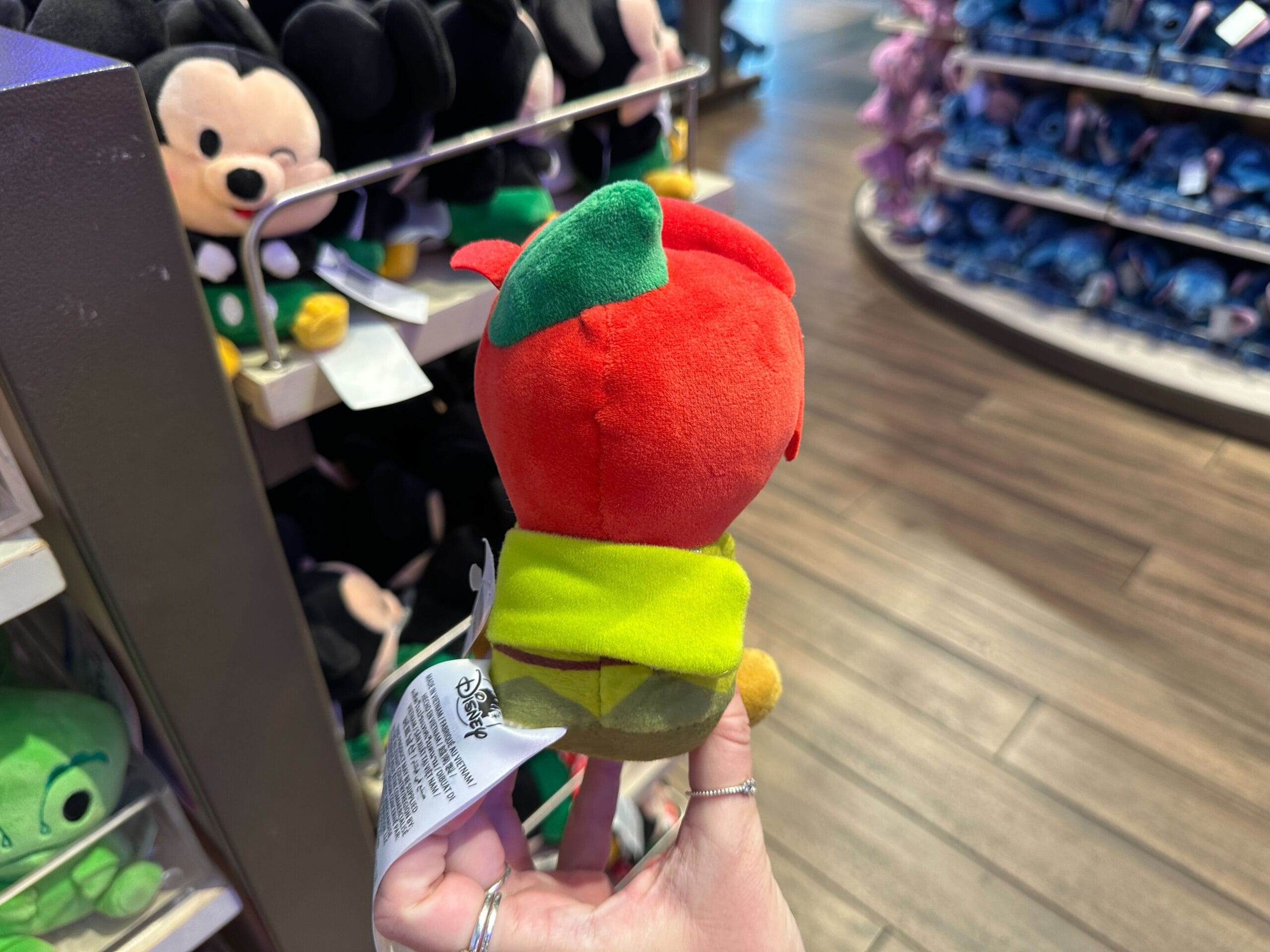 A hand holds a red and green plush toy in a store aisle lined with Disney character plushies, reminiscent of Main Street shops.