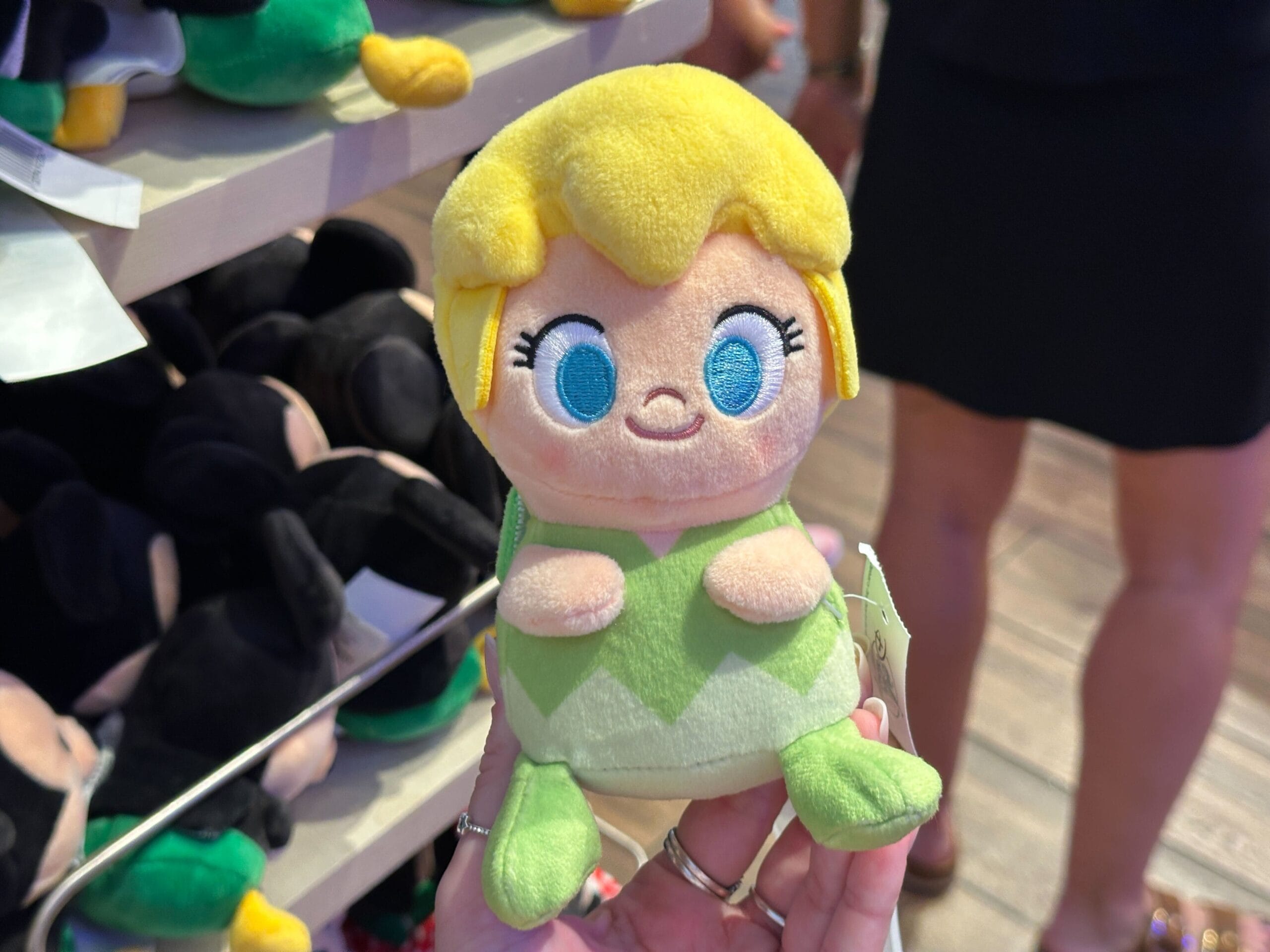 A hand holds a small, plush Tinker Bell doll in a Disney park store, with other plush toys displayed on shelves behind it.