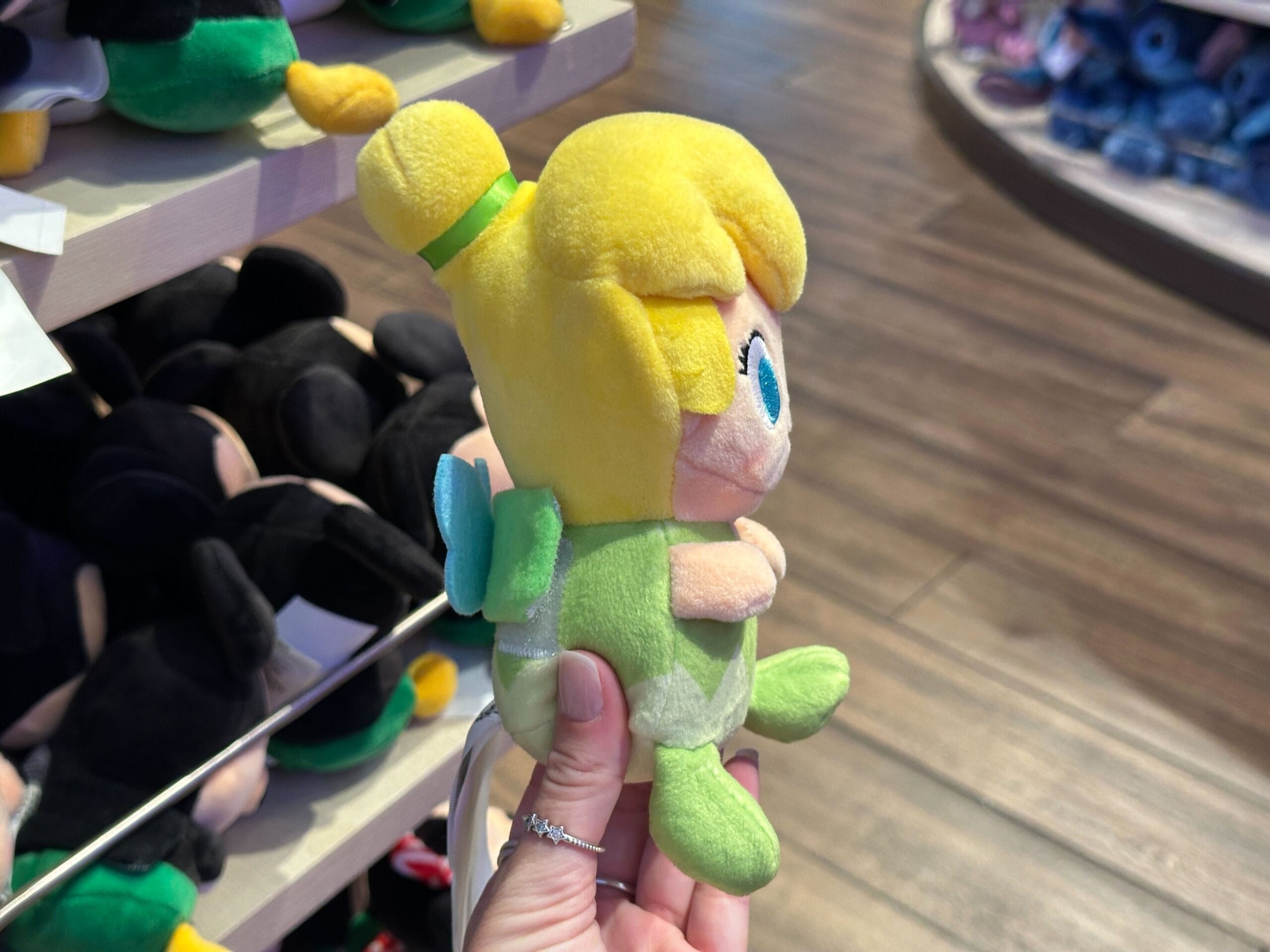A hand holds a Tinker Bell plush toy in a Disney park gift shop, surrounded by other plush toys displayed on nearby shelves.