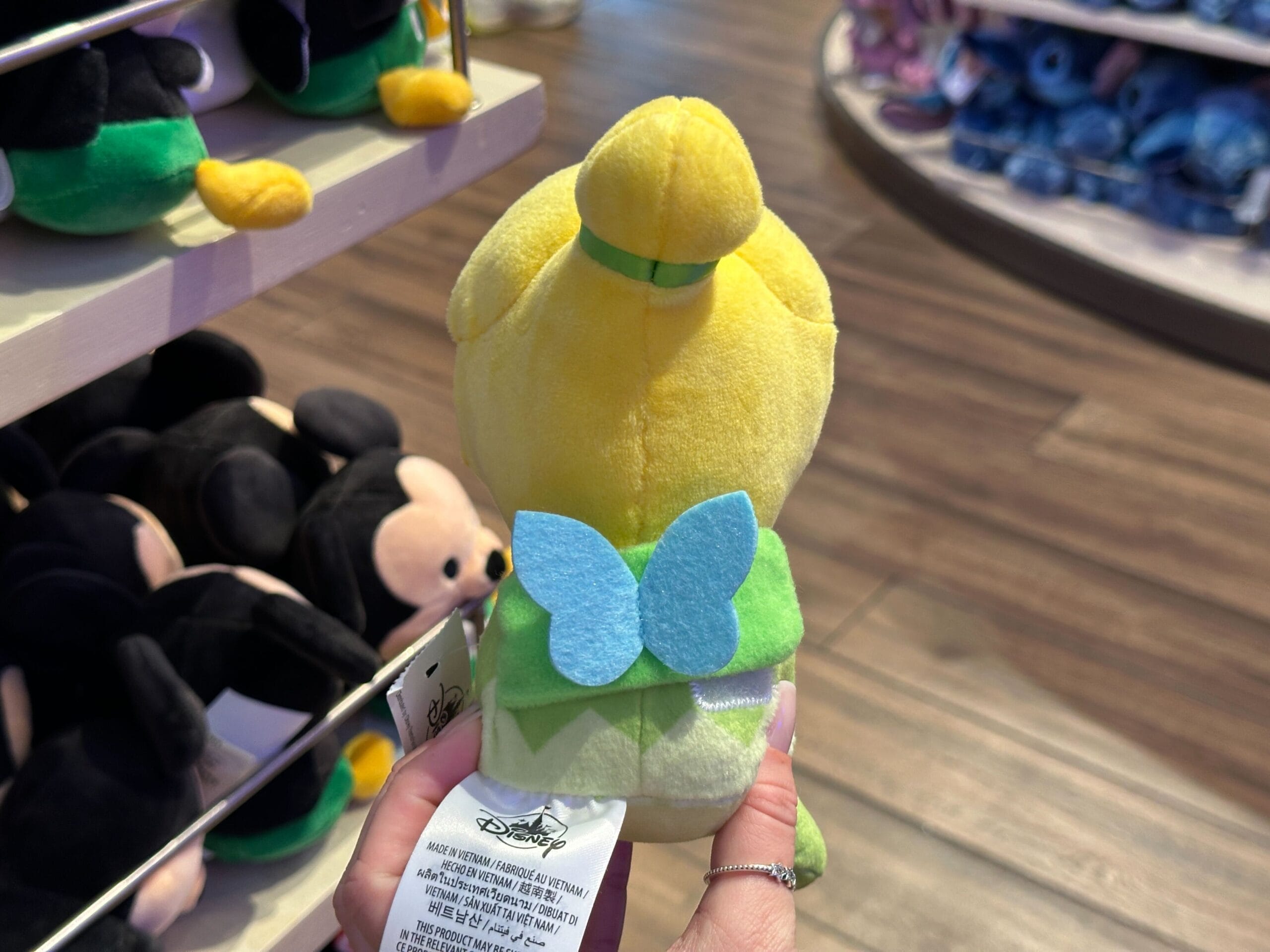 A hand holds a Tinker Bell plush with blue wings in a Disney park shop, surrounded by stuffed characters from classic Disney films.