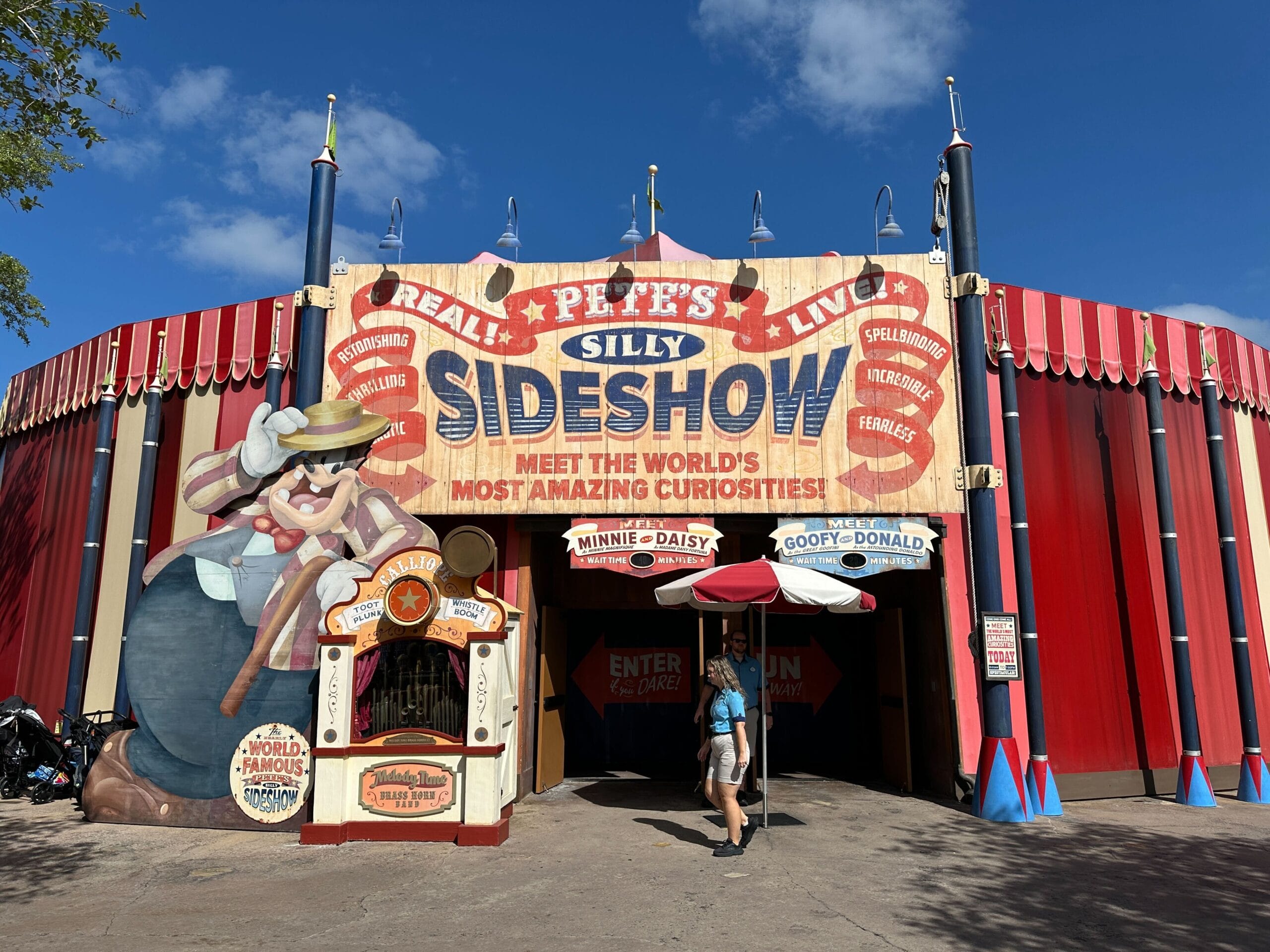 A woman walks into Pete’s Silly Sideshow circus tent attraction with colorful cartoon signs under a blue sky.