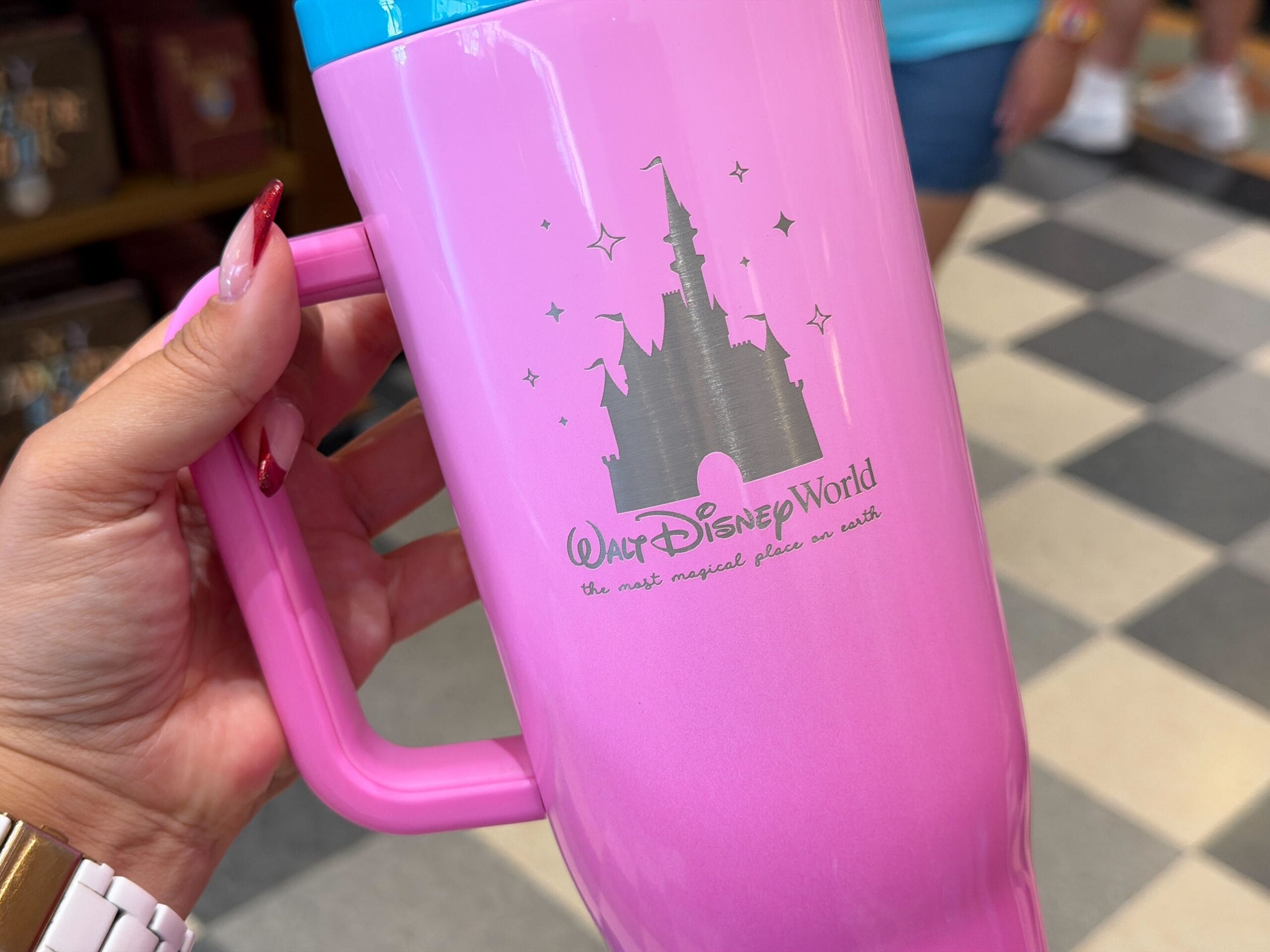 A hand holds a pink Walt Disney World mug featuring Cinderella Castle—distinct from Sleeping Beauty or Shanghai castles—in a store with checkered floor.