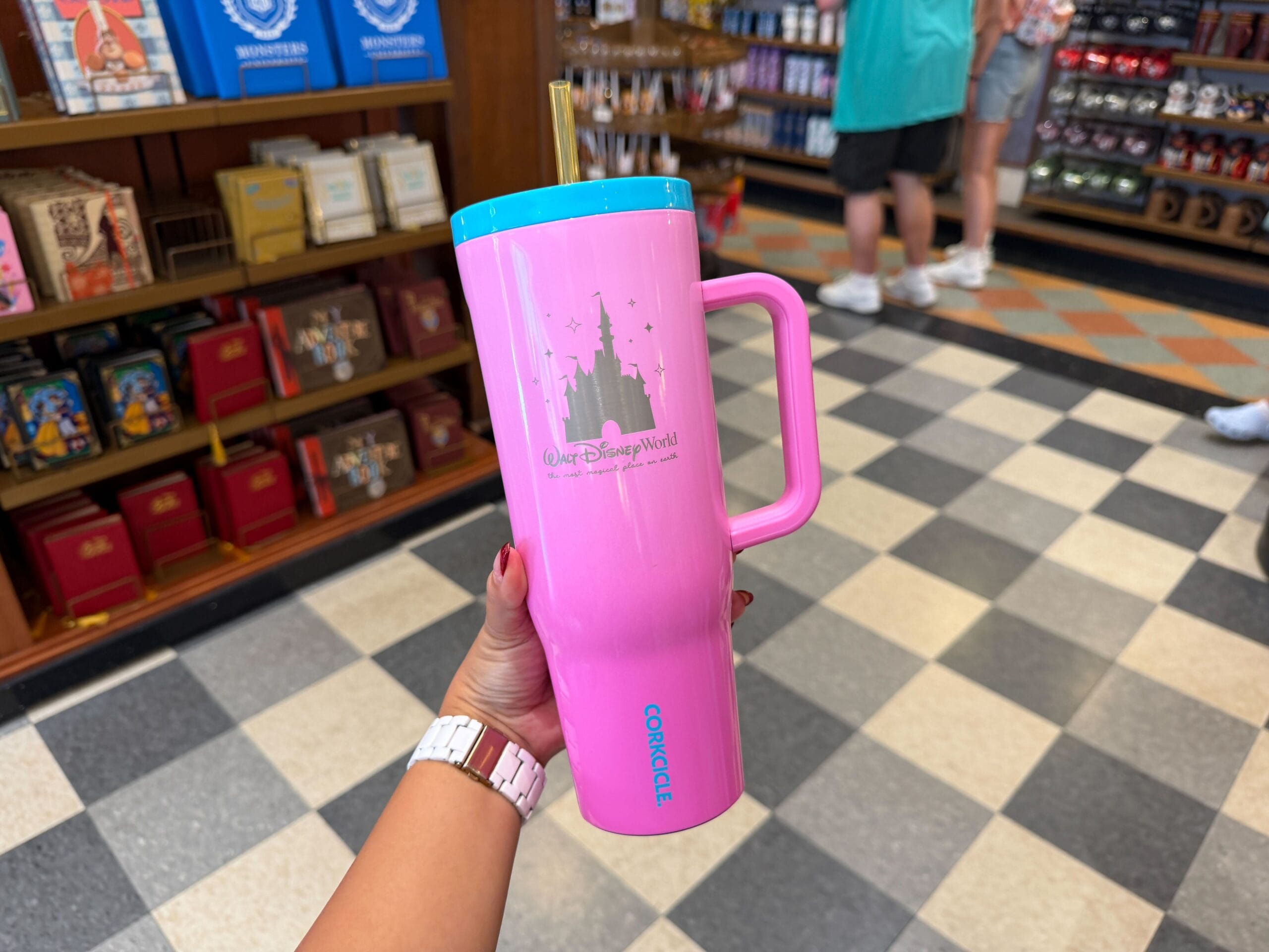 A hand holds a large pink Walt Disney World Corkcicle tumbler inside a park shop with checkered floors, reminiscent of Main Street U.S.A.