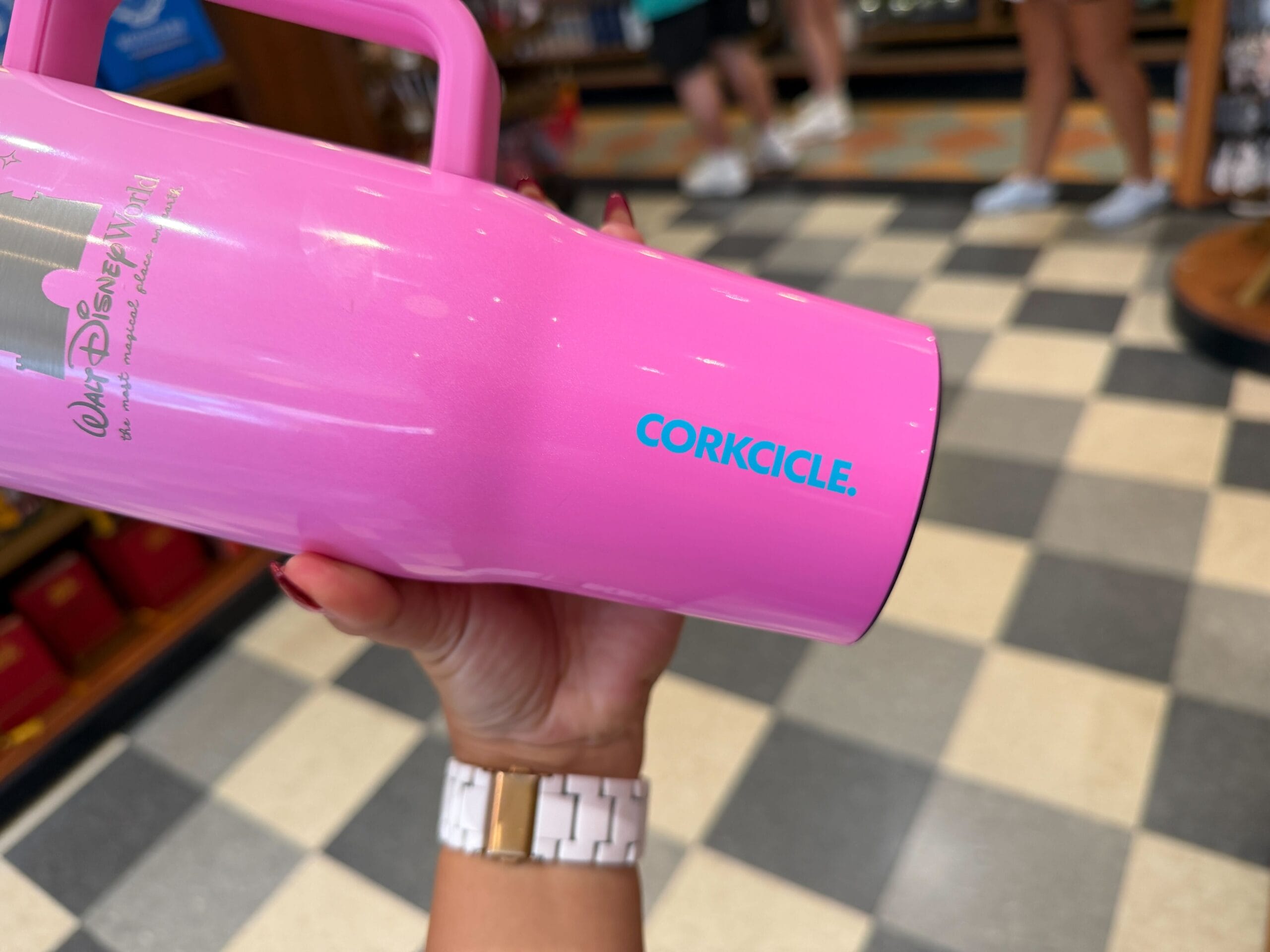 A hand holds a pink Corkcicle cup inside a Disney park shop with a black and white checkered floor, vintage boutique vibes.
