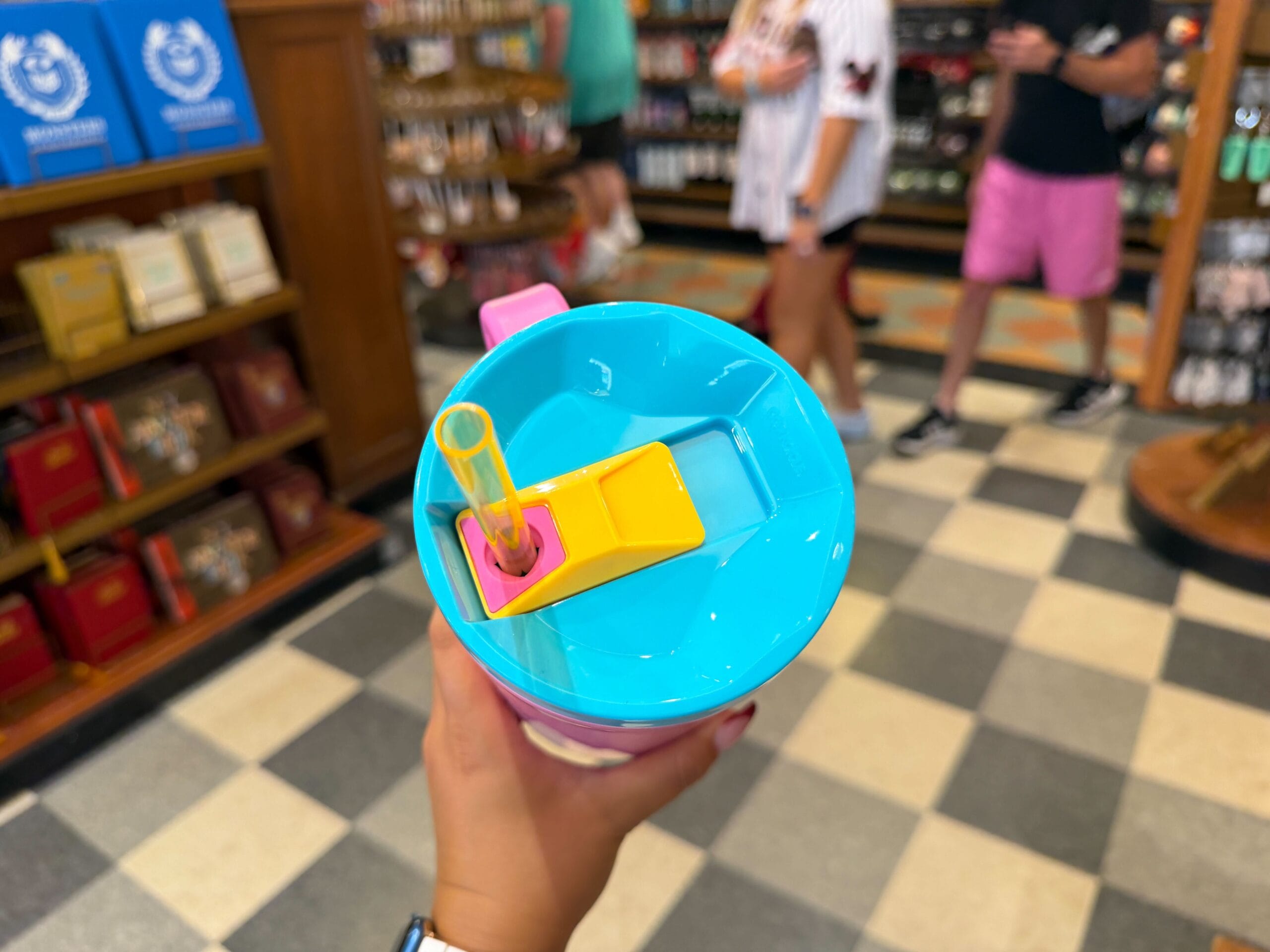 A hand holds a blue cup with yellow lid and two straws in a Disney park shop, with guests and merch shelves in the background.