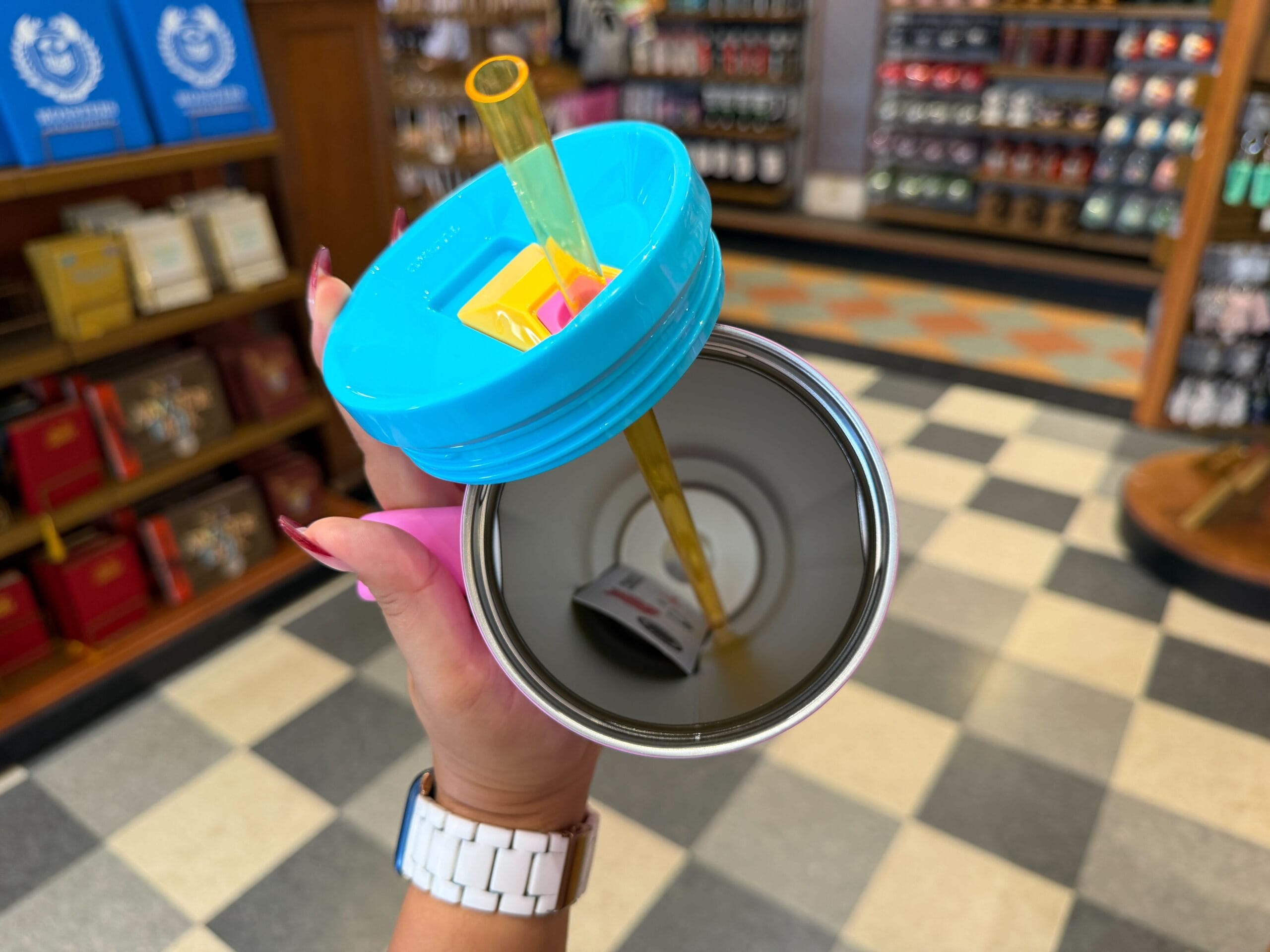 A hand holds an open tumbler with a blue lid and yellow straw inside a Disney park shop with checkered flooring.