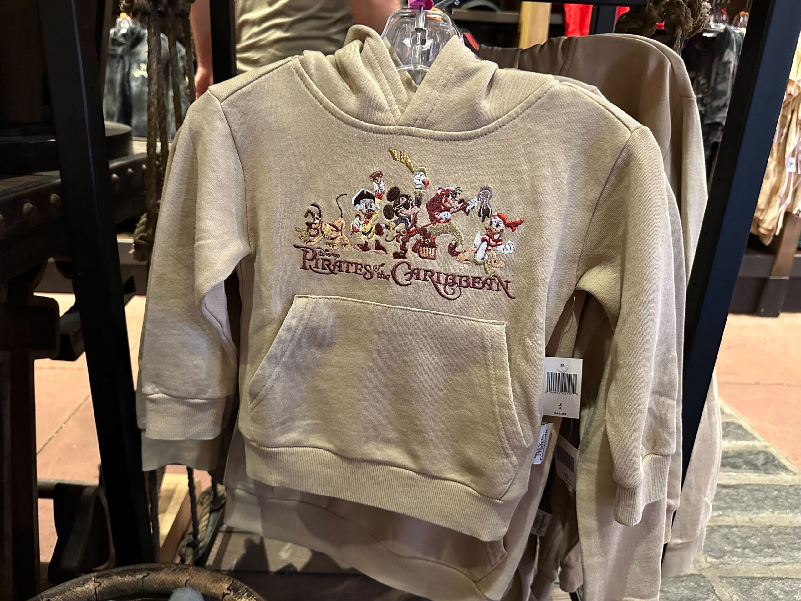 Beige Pirates of the Caribbean hoodie with embroidered cartoon pirate crew, displayed on a rack at a Disney park merchandise shop.