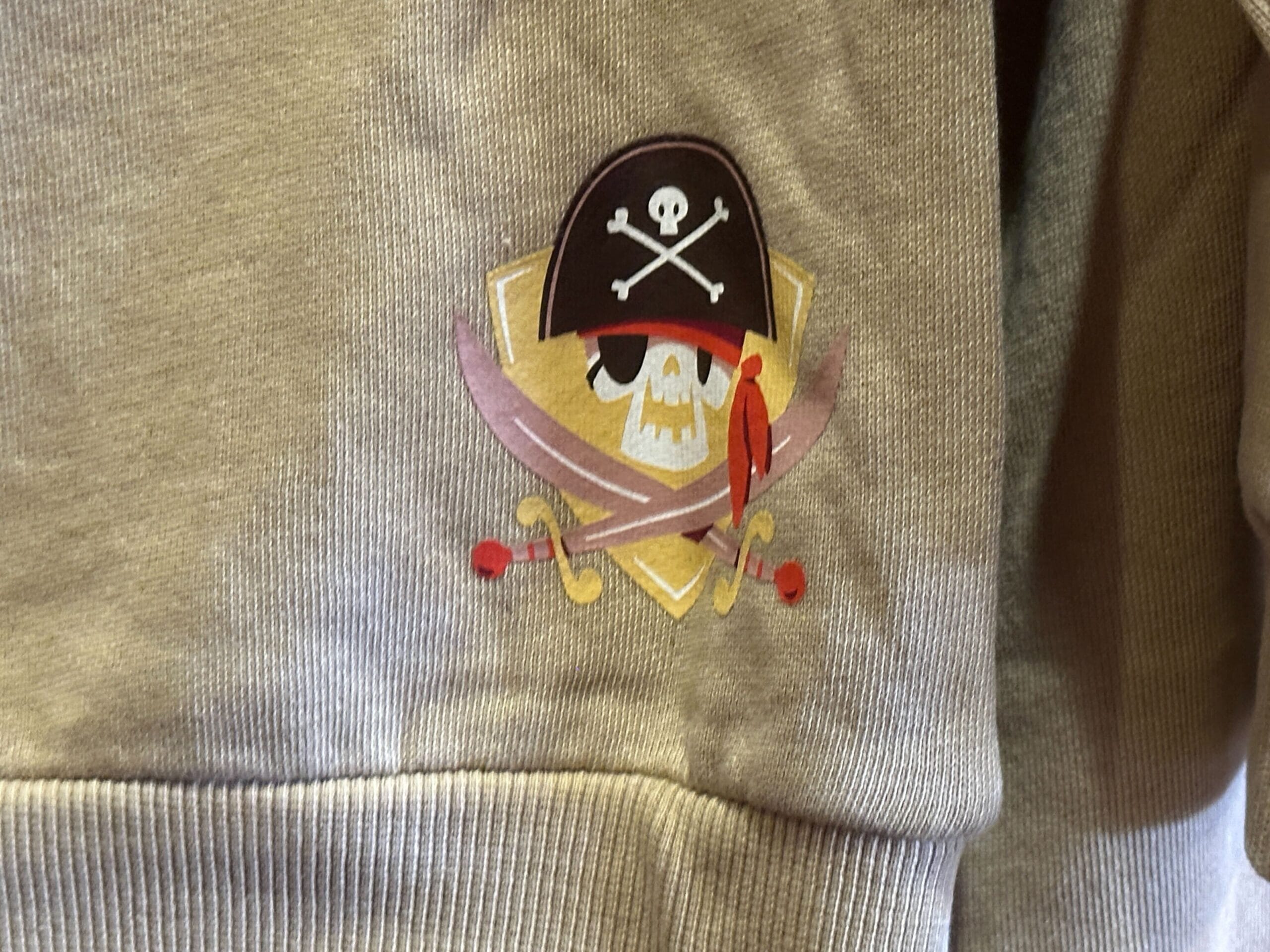 A whimsical cartoon pirate skull with eye patch, hat, and crossed swords, set on a tan background reminiscent of Disney park themes.