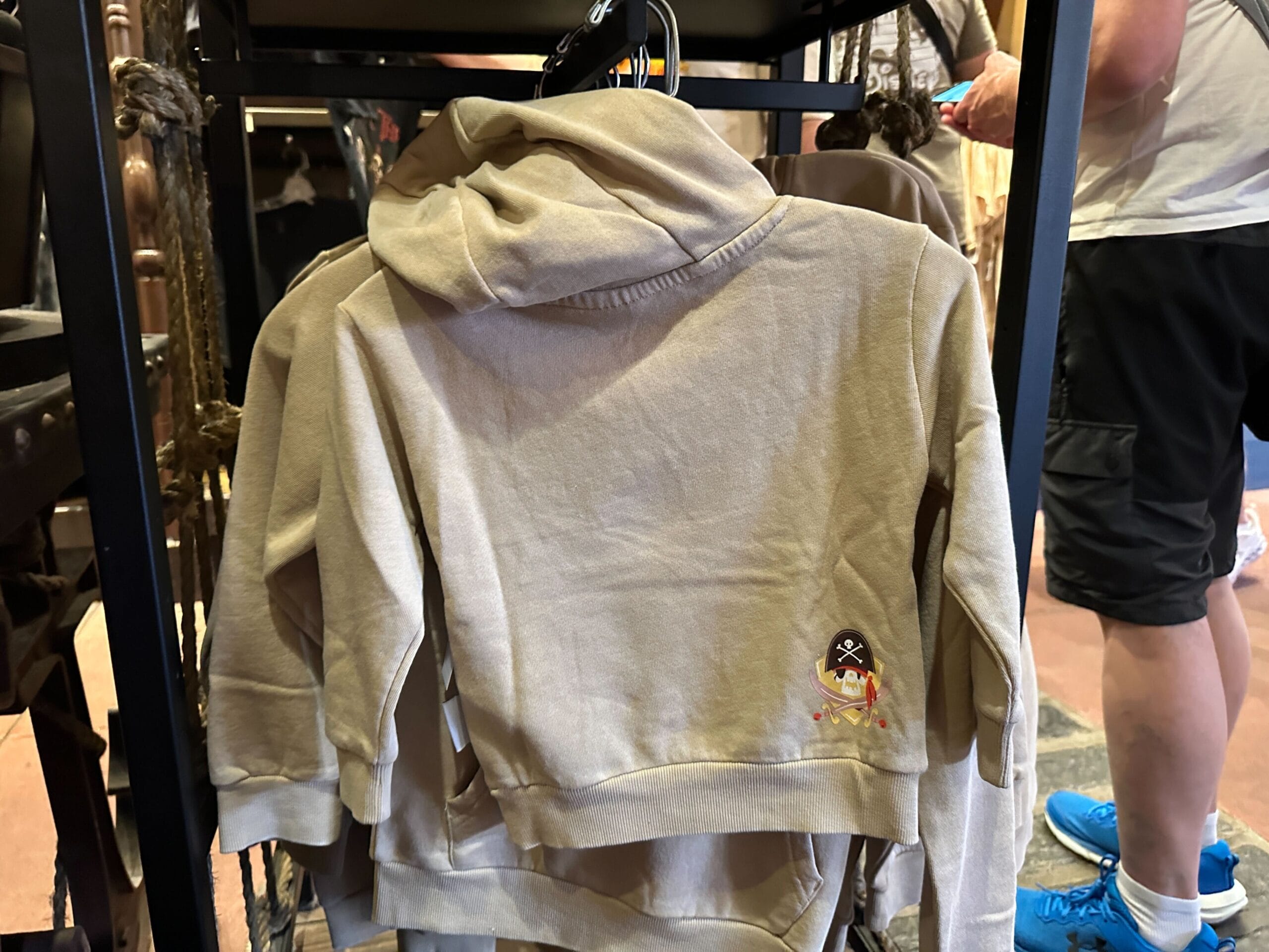 Tan hoodie featuring a small pirate face and crossed swords on the lower back, hanging on a store rack at a Disney Park shop.