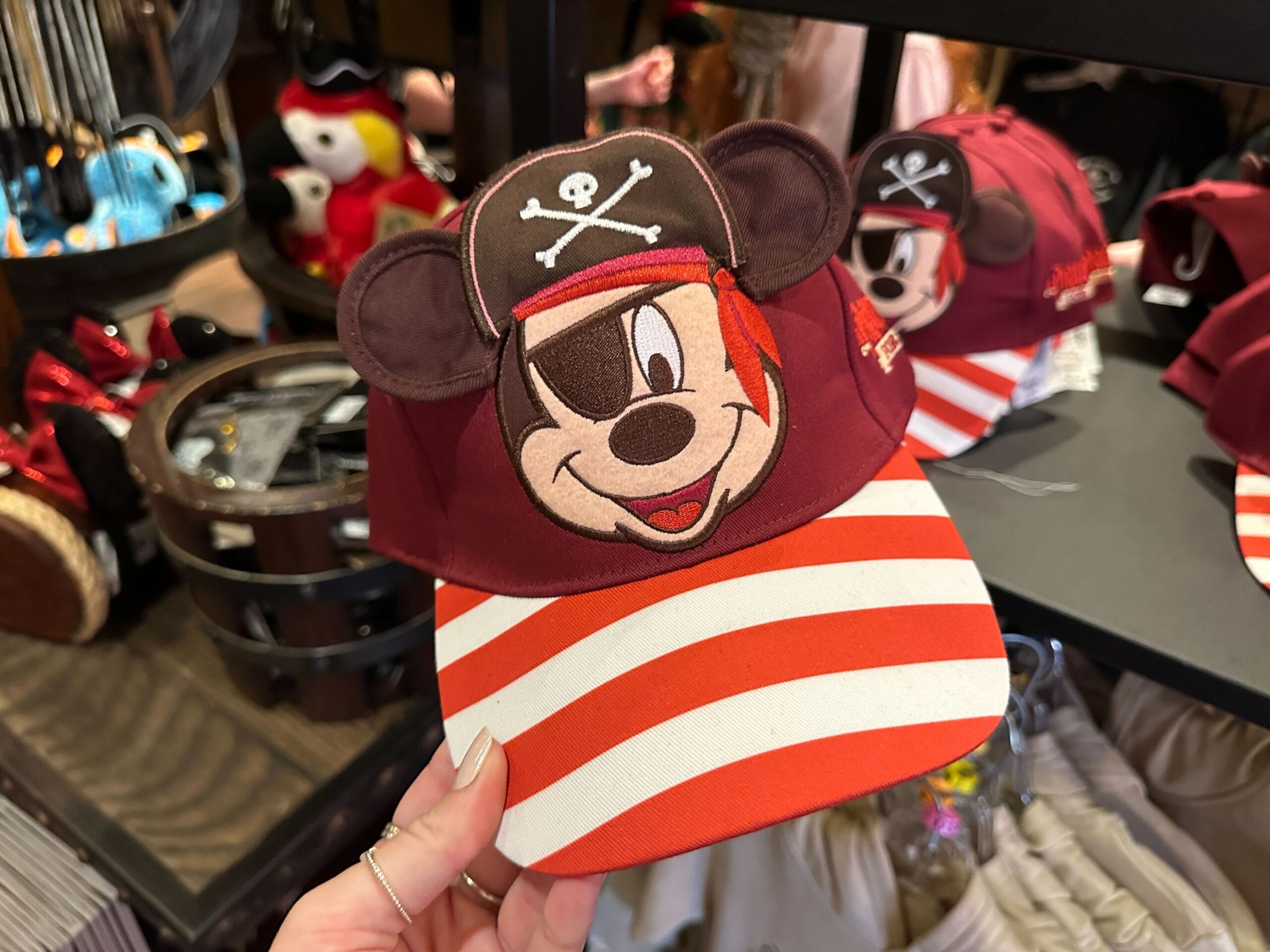 A hand holds a pirate-themed Mickey Mouse hat with ears and a red-and-white striped brim, perfect for Disney Park adventures.