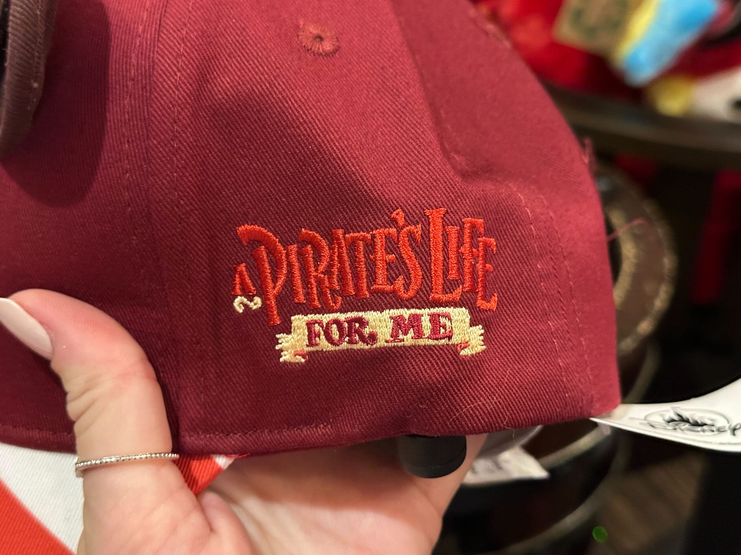 A hand holds a red cap featuring "A Pirate's Life For Me" in red and gold thread, reminiscent of Disney Parks' pirate themes.