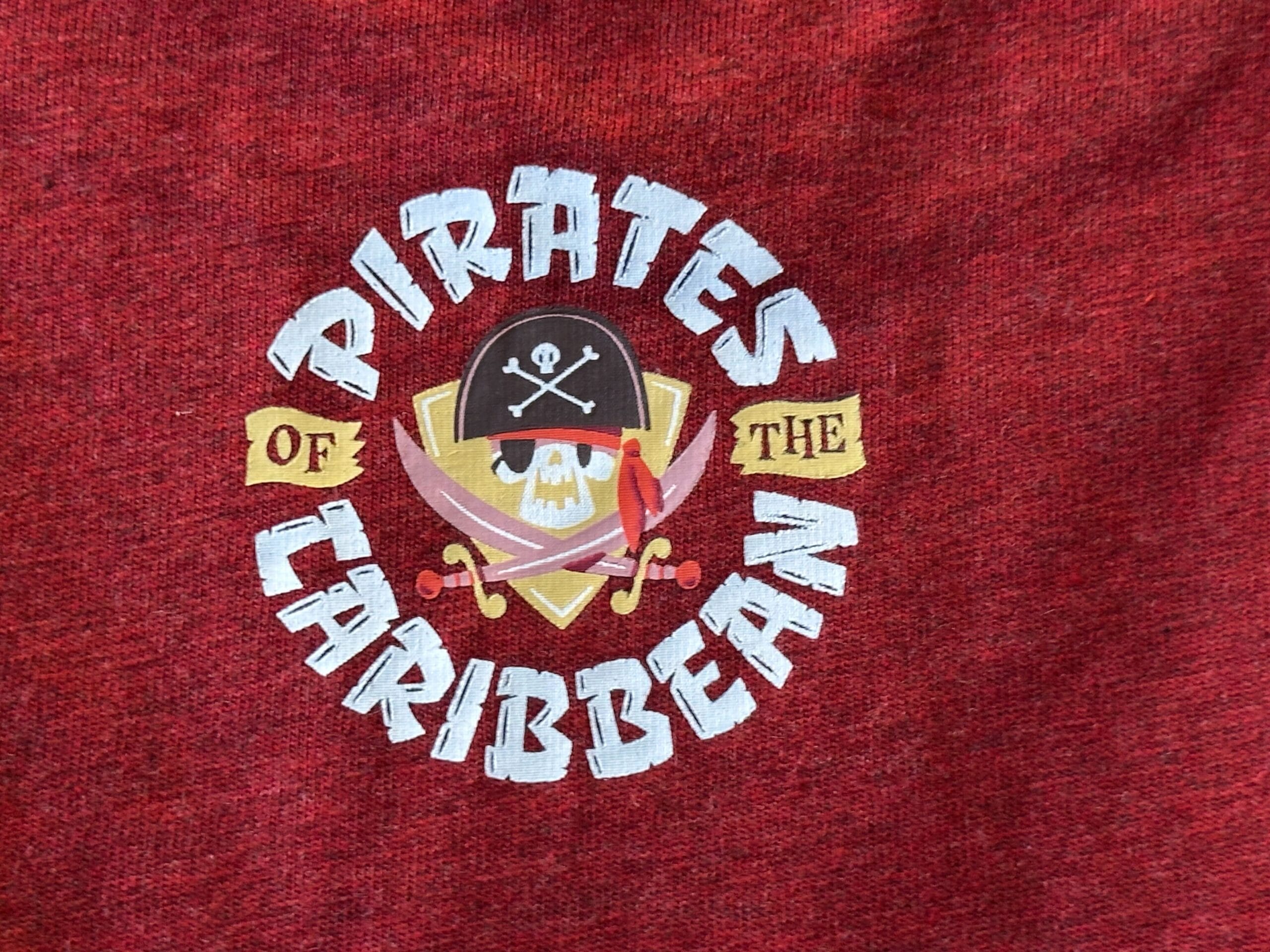 Pirates of the Caribbean logo with pirate skull, crossed swords, and shield on rich red fabric—iconic in many Disney Parks worldwide.