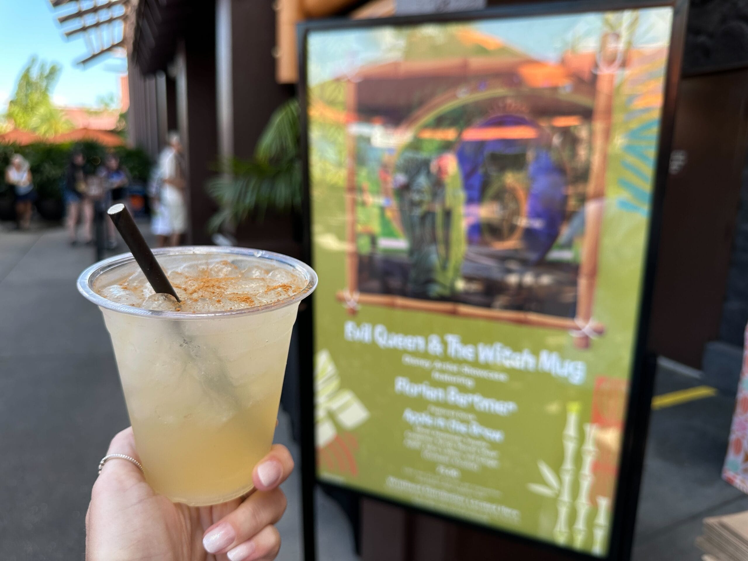 Hand holding a plastic cup with a light drink at Disney's Polynesian Village Resort, colorful event sign outside, no castle visible.