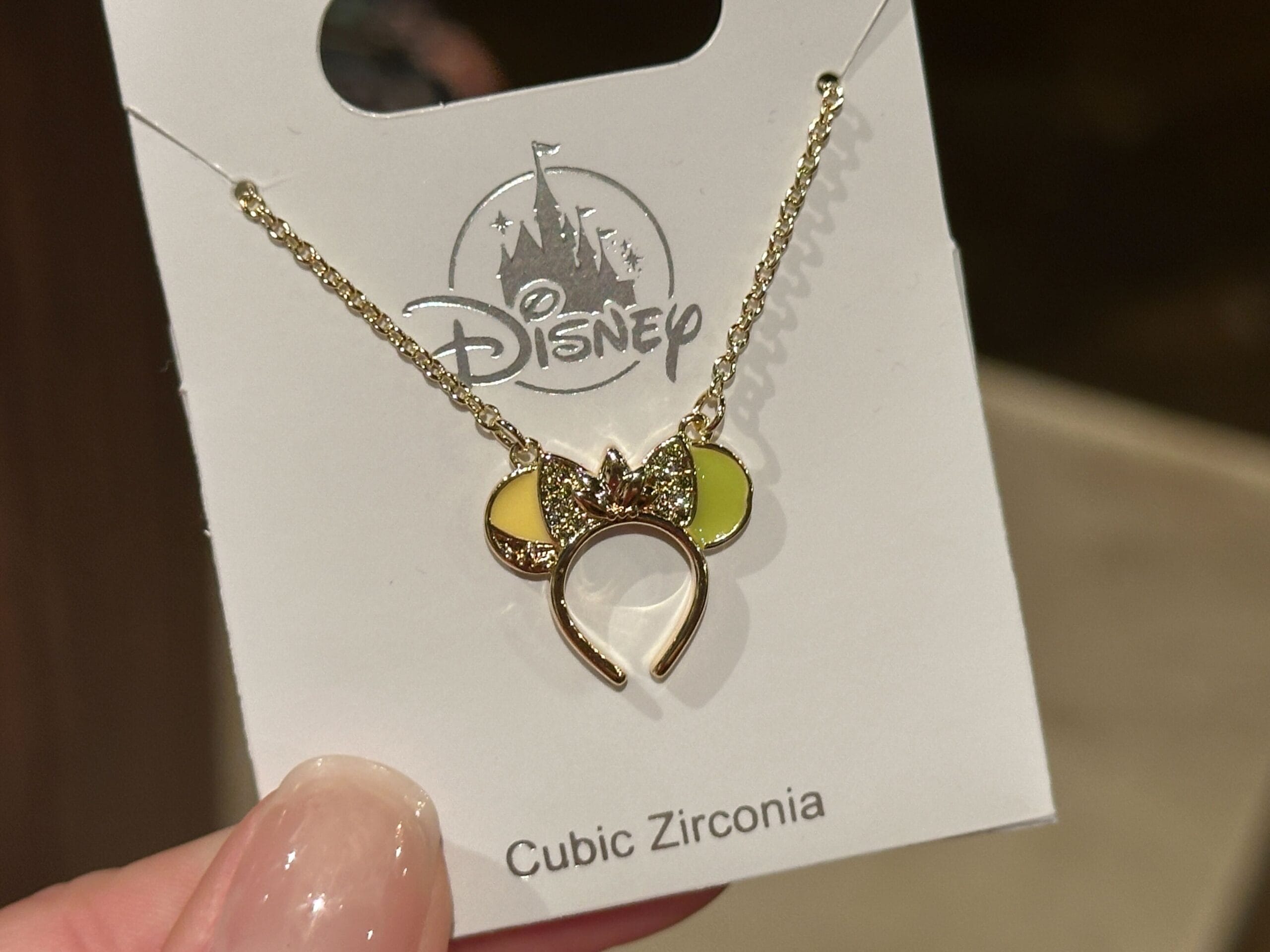 Gold necklace with Minnie Mouse ears pendant, yellow accents and bow, on a Disney card marked "Cubic Zirconia," evoking park charm.