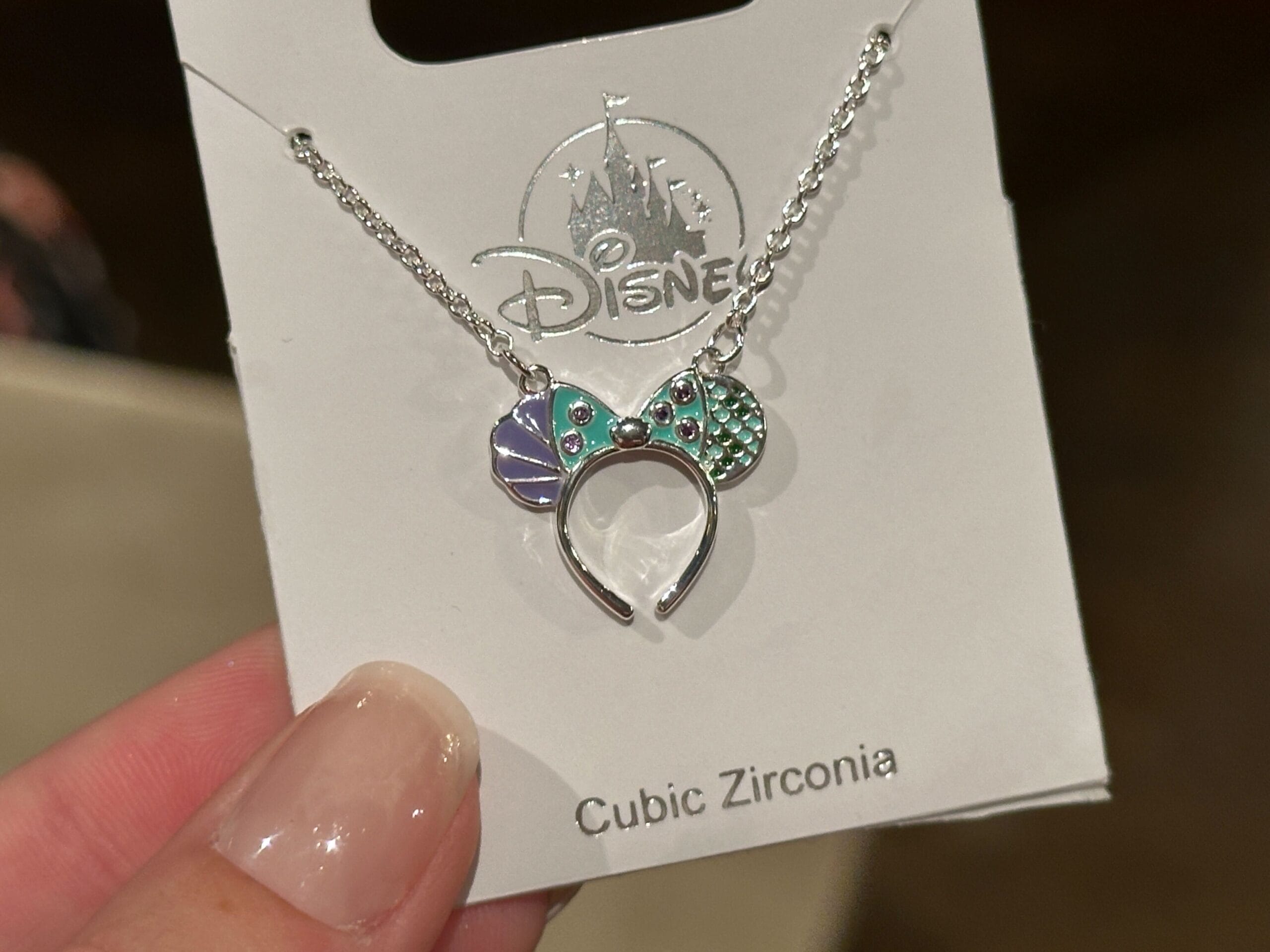 A hand holds a Disney necklace featuring Minnie Mouse ears in purple, green, and blue, displayed on card—typical of park merch.