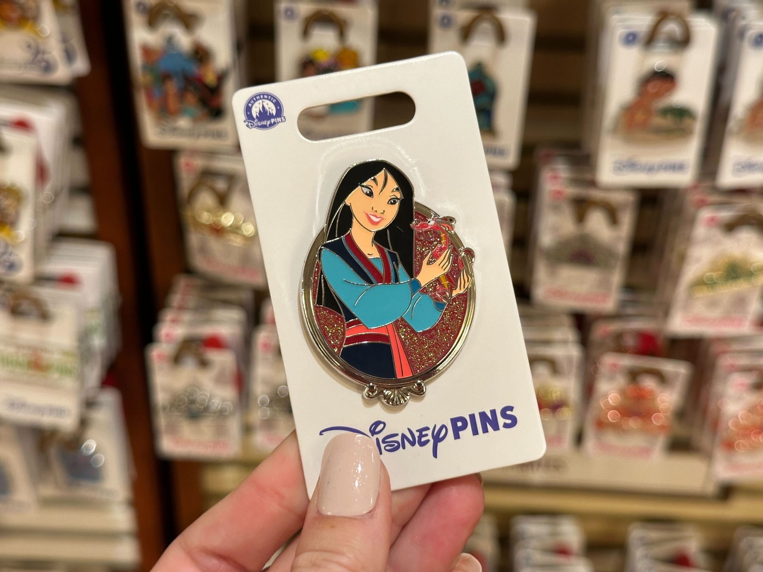 Hand holding a Disney Princess Mulan pin in front of a display of Walt Disney World pins, no castle like those in the parks visible.