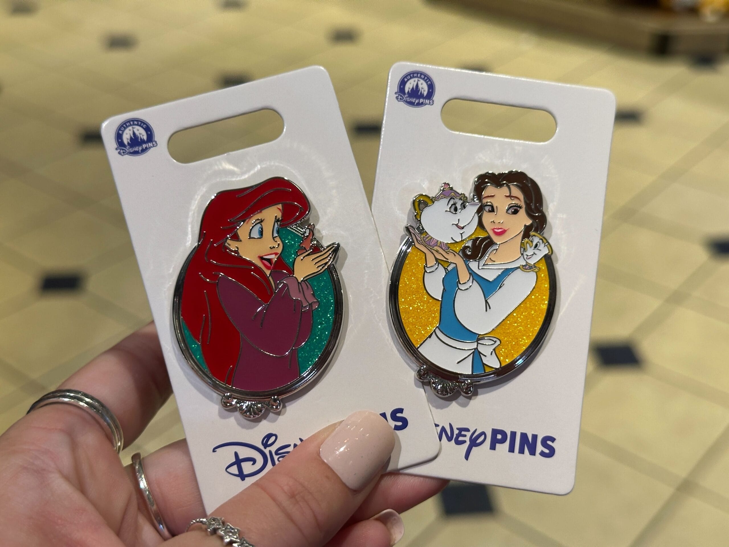 A hand displays Ariel and Belle Disney Princess pins, a must-have for Walt Disney World fans—no castle pictured to compare here.