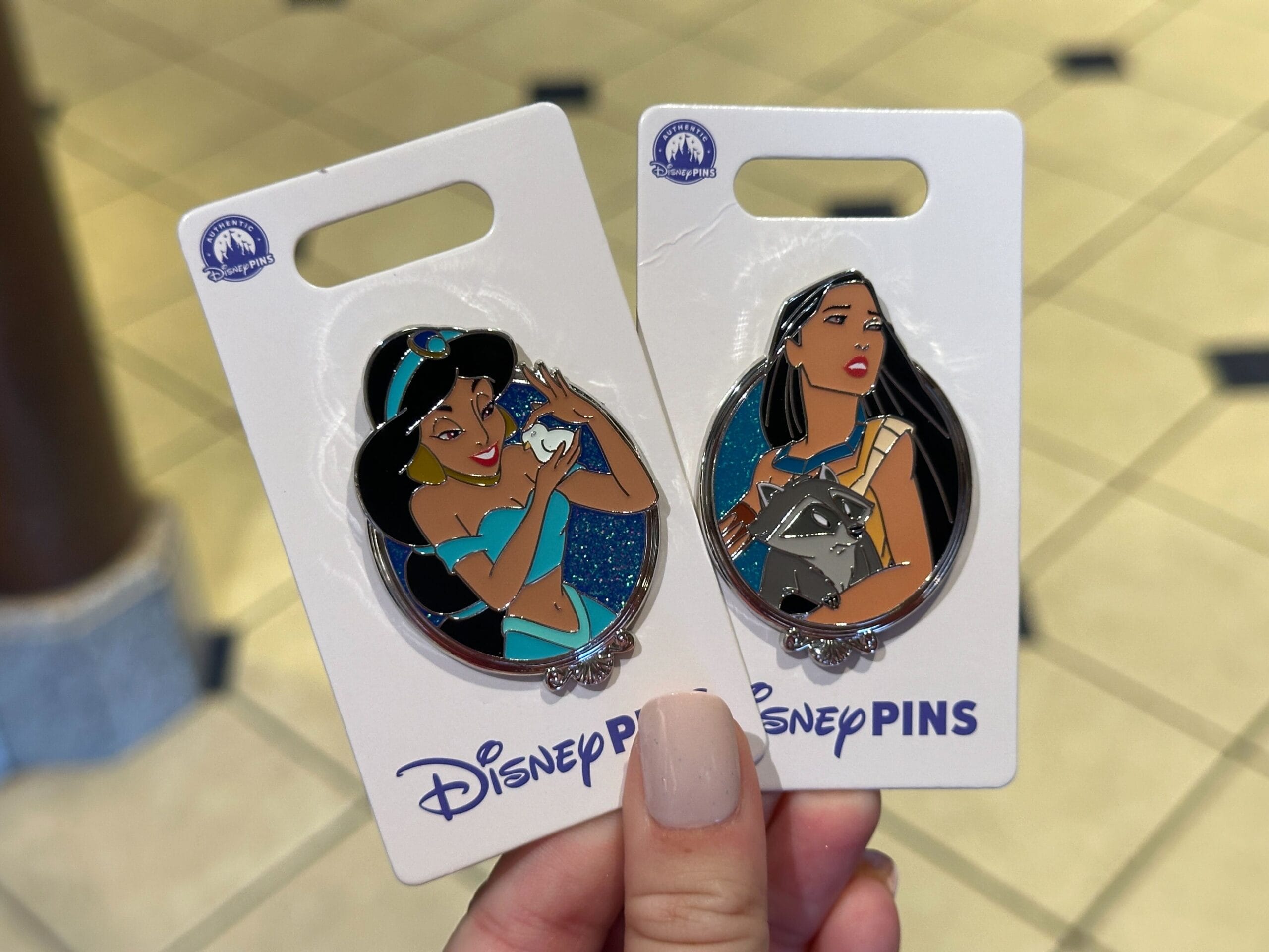 A hand displays two Walt Disney World pins of Princess Jasmine and Pocahontas on white backing cards, no castles pictured here.