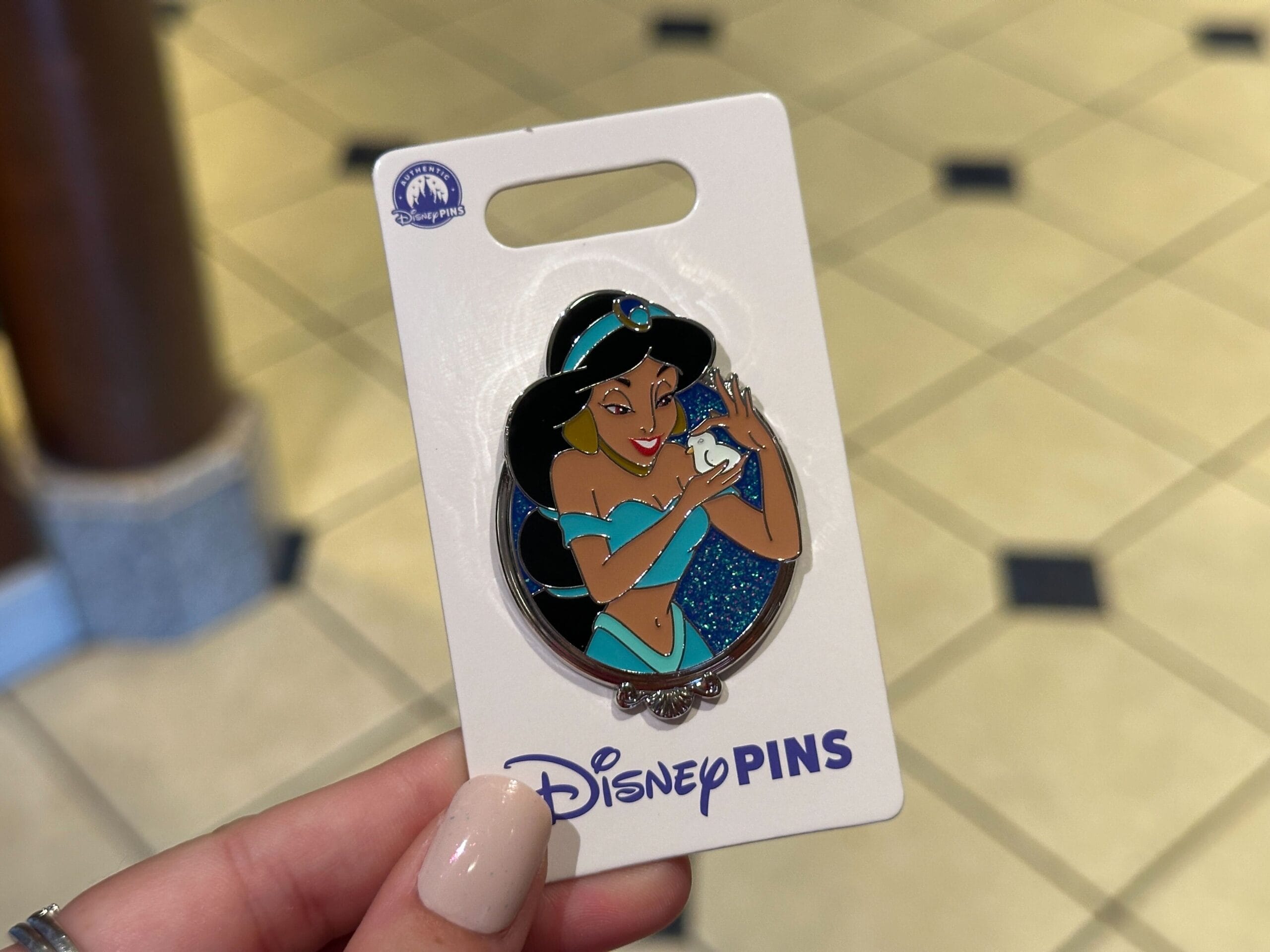 A hand displays a Jasmine pin from Aladdin on a "Disney Pins" card at Walt Disney World, home to Cinderella Castle.