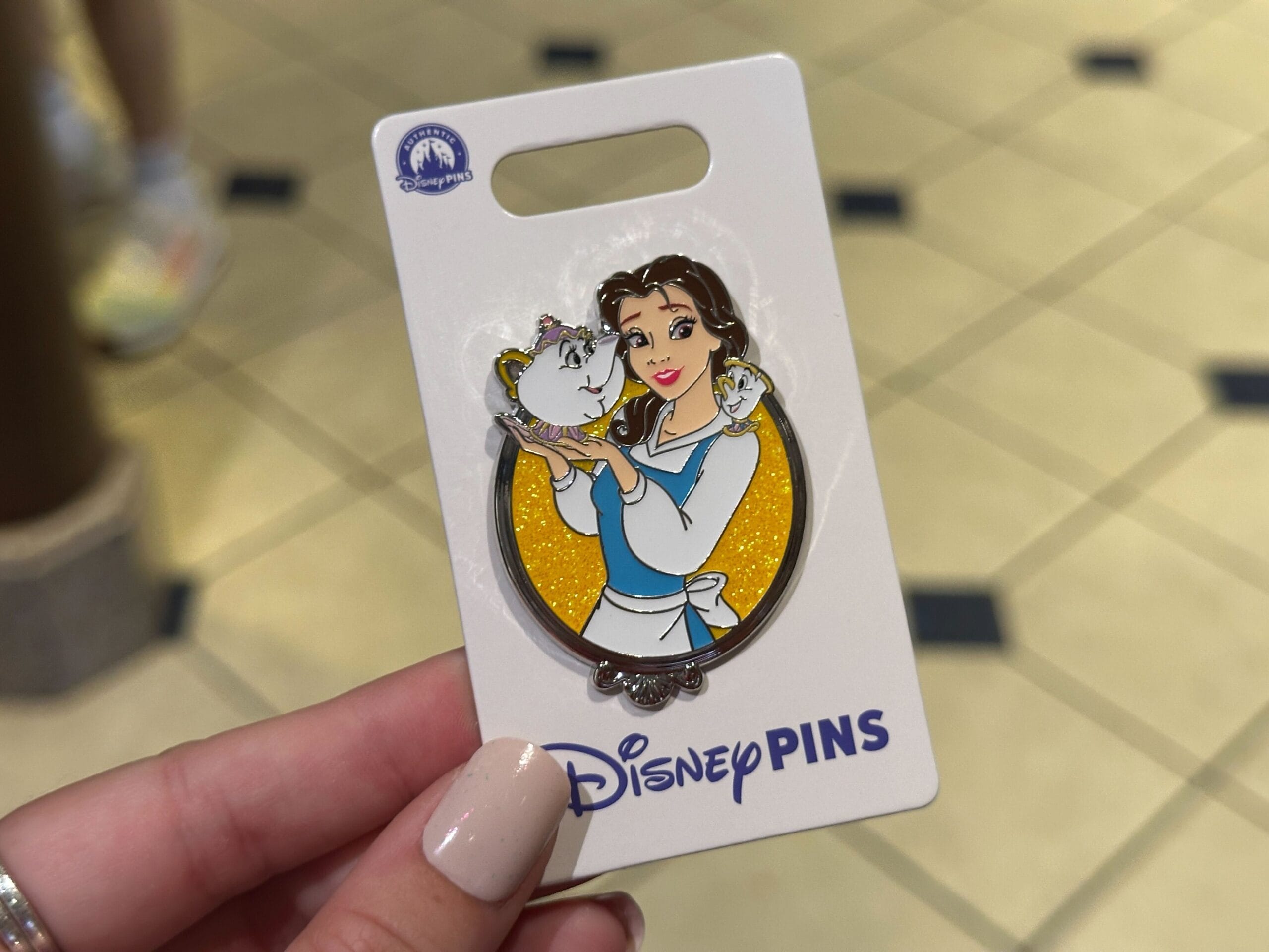 A hand at a Disney park displays a Princess Sidekicks Pin with Belle, Mrs. Potts, and Chip—no castle backdrop in this shot.