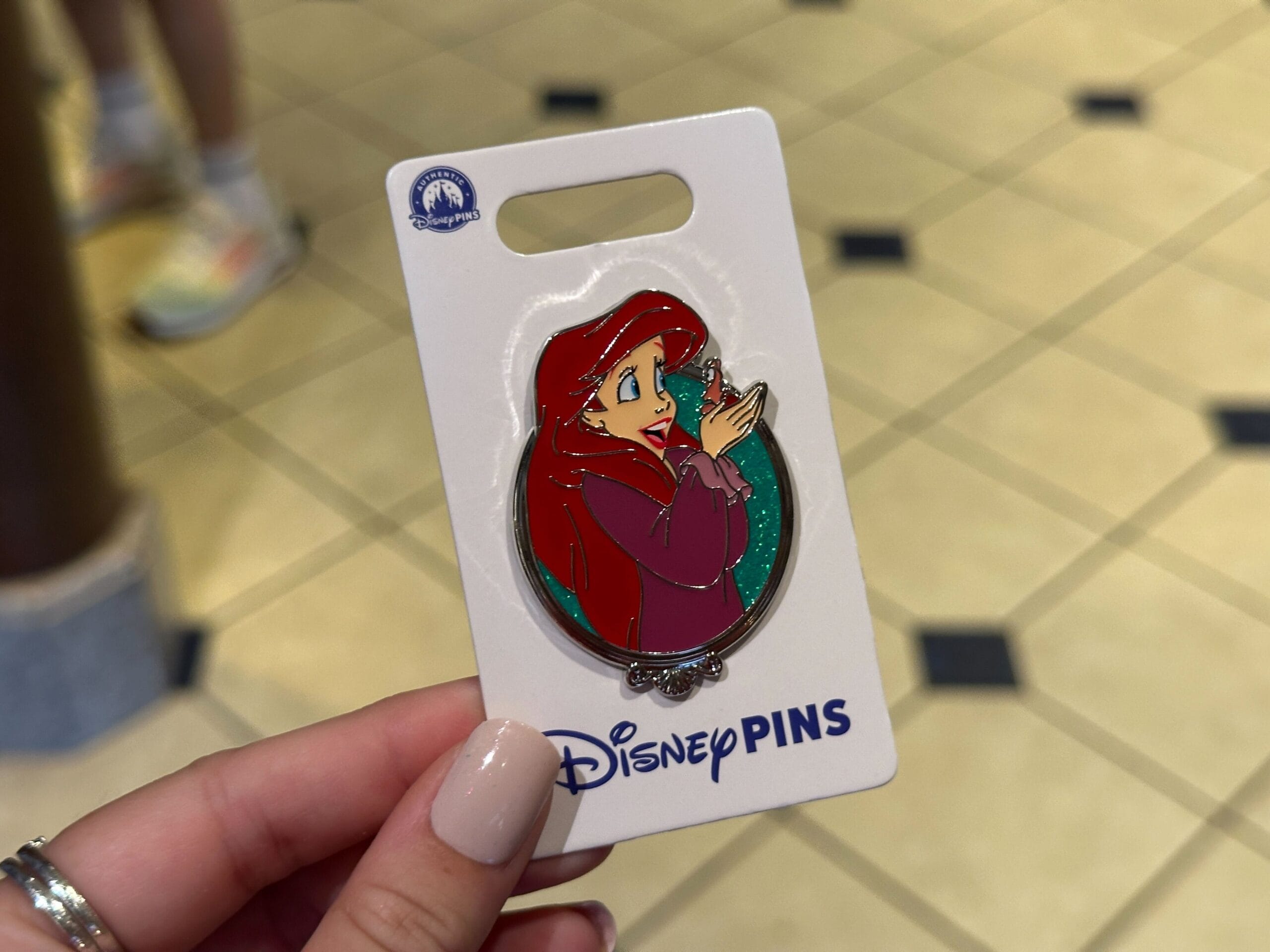 A hand displays an Ariel pin from The Little Mermaid on a tiled floor at Walt Disney World, home to Cinderella Castle, not Sleeping Beauty Castle.