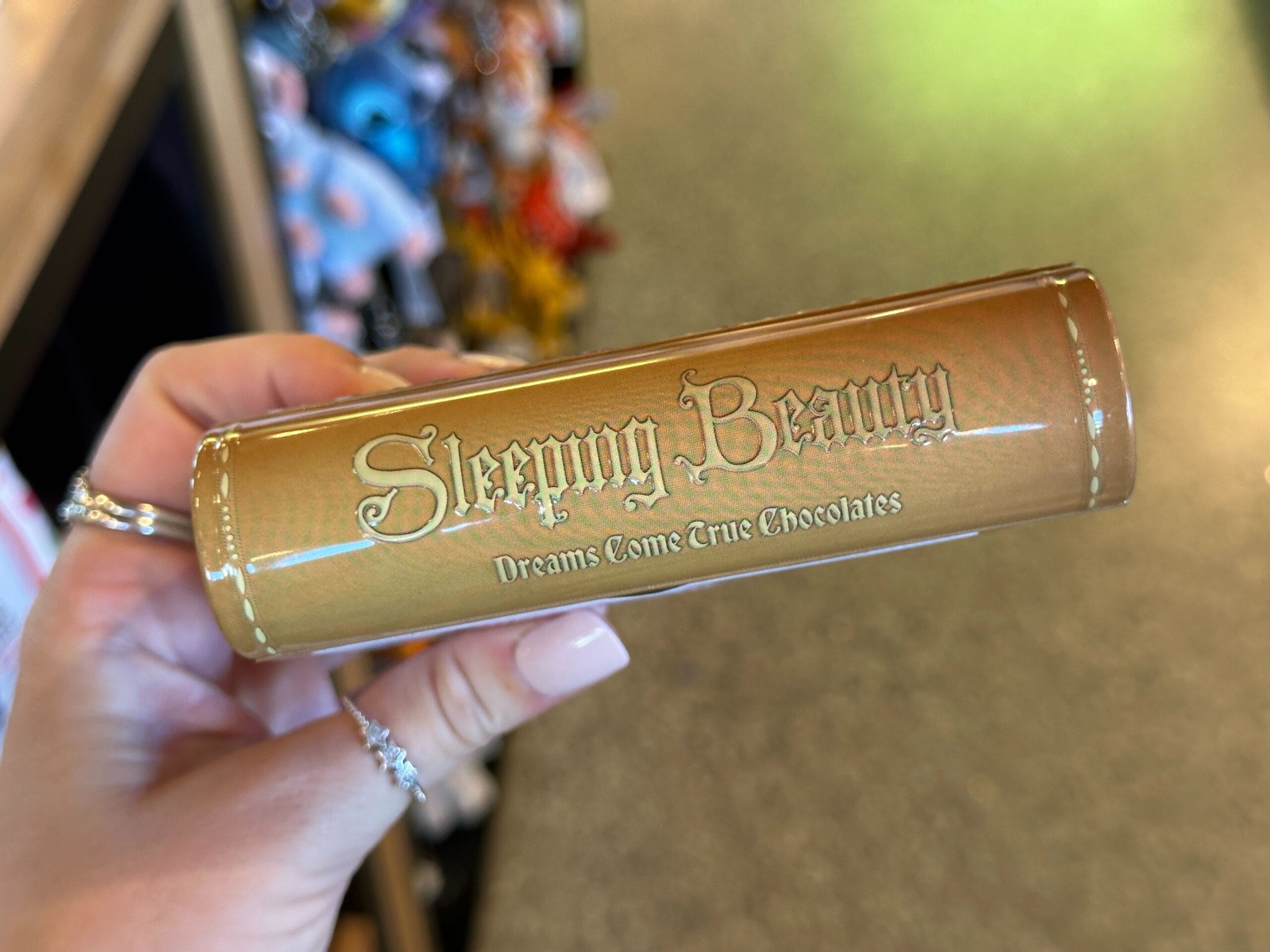 A hand holds a gold tube labeled "Sleeping Beauty Dreams Come True Chocolates," inspired by the Disneyland castle’s fairytale charm.