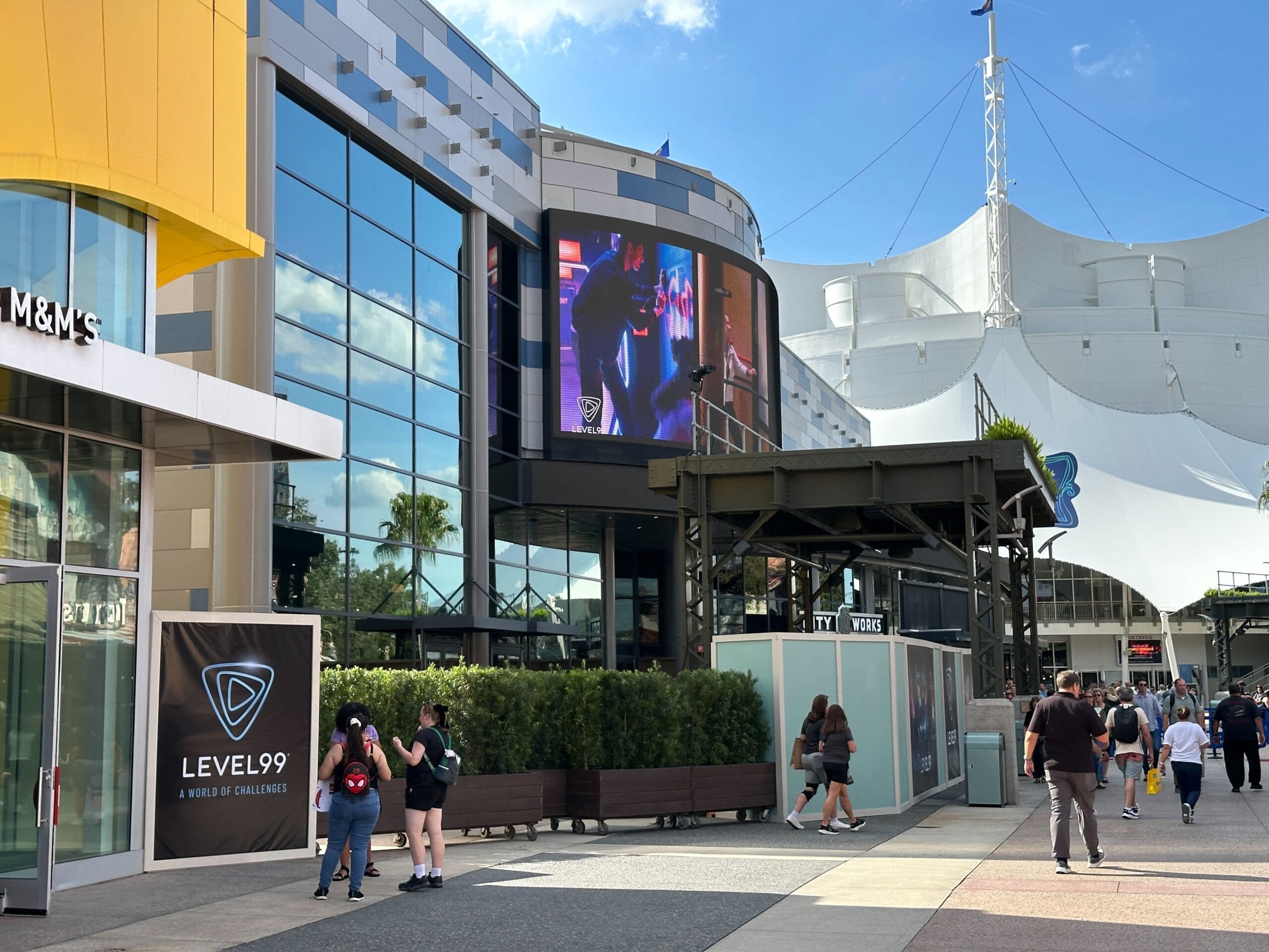 Visitors stroll by themed shops and a large screen at an outdoor plaza—no castle in sight like Disneyland or Magic Kingdom.