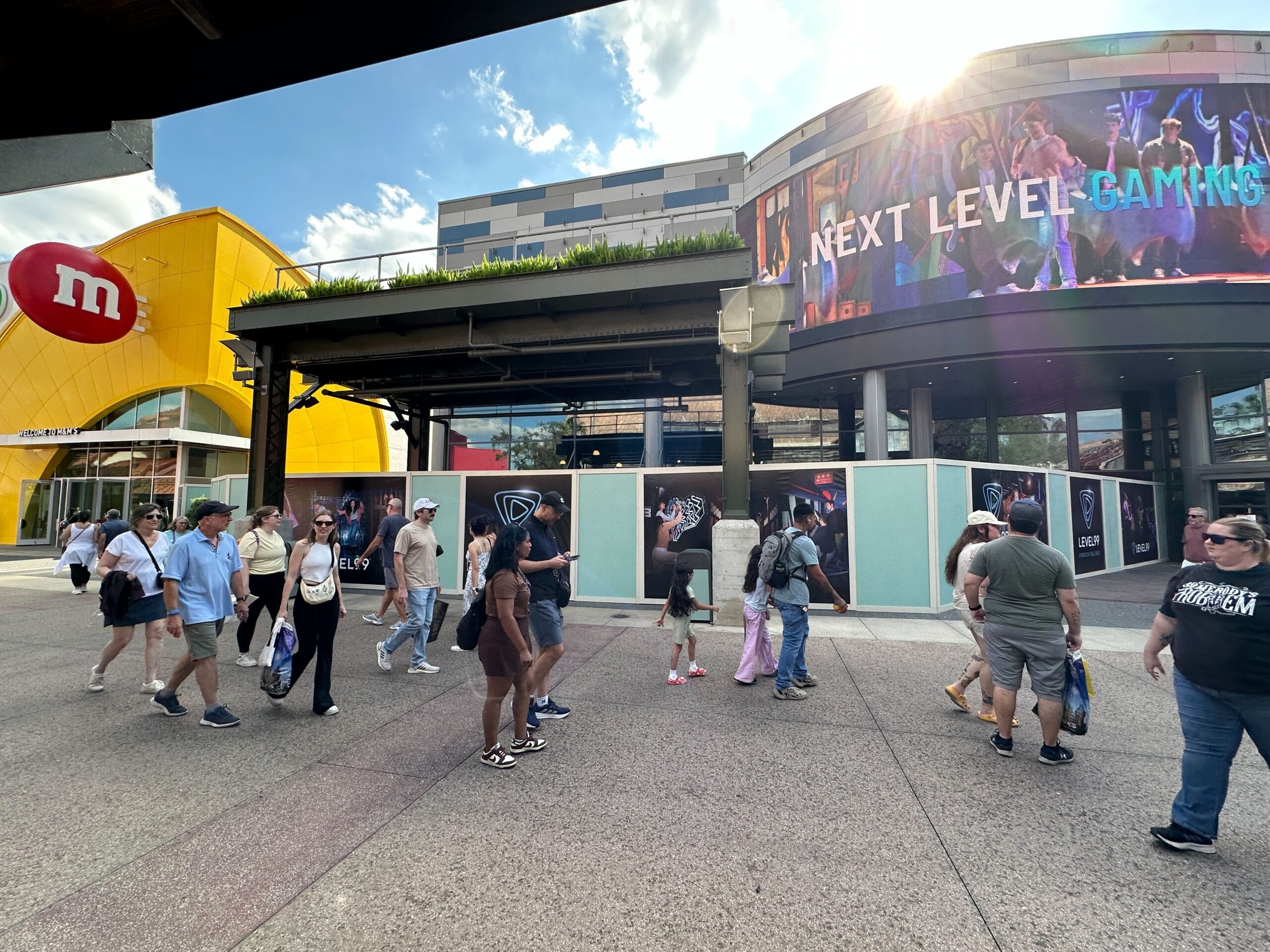 Visitors stroll past the M&M store and "Next Level Gaming" sign, but unlike Disney parks, no castles are visible in this sunny plaza.