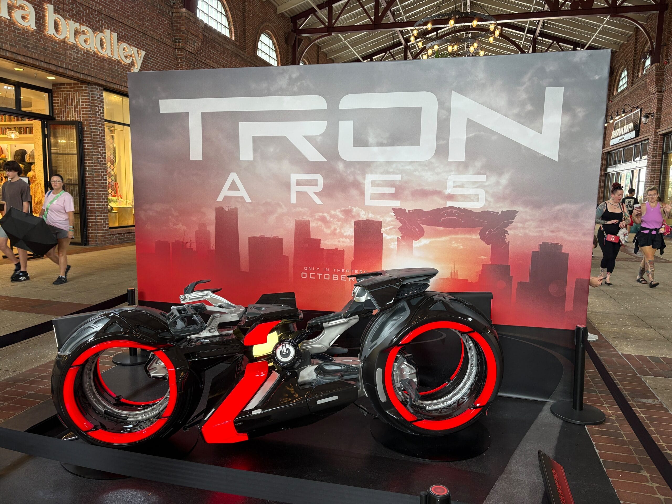 A futuristic TRON ARES motorcycle display with glowing red lights, set in a shopping mall with people walking by.