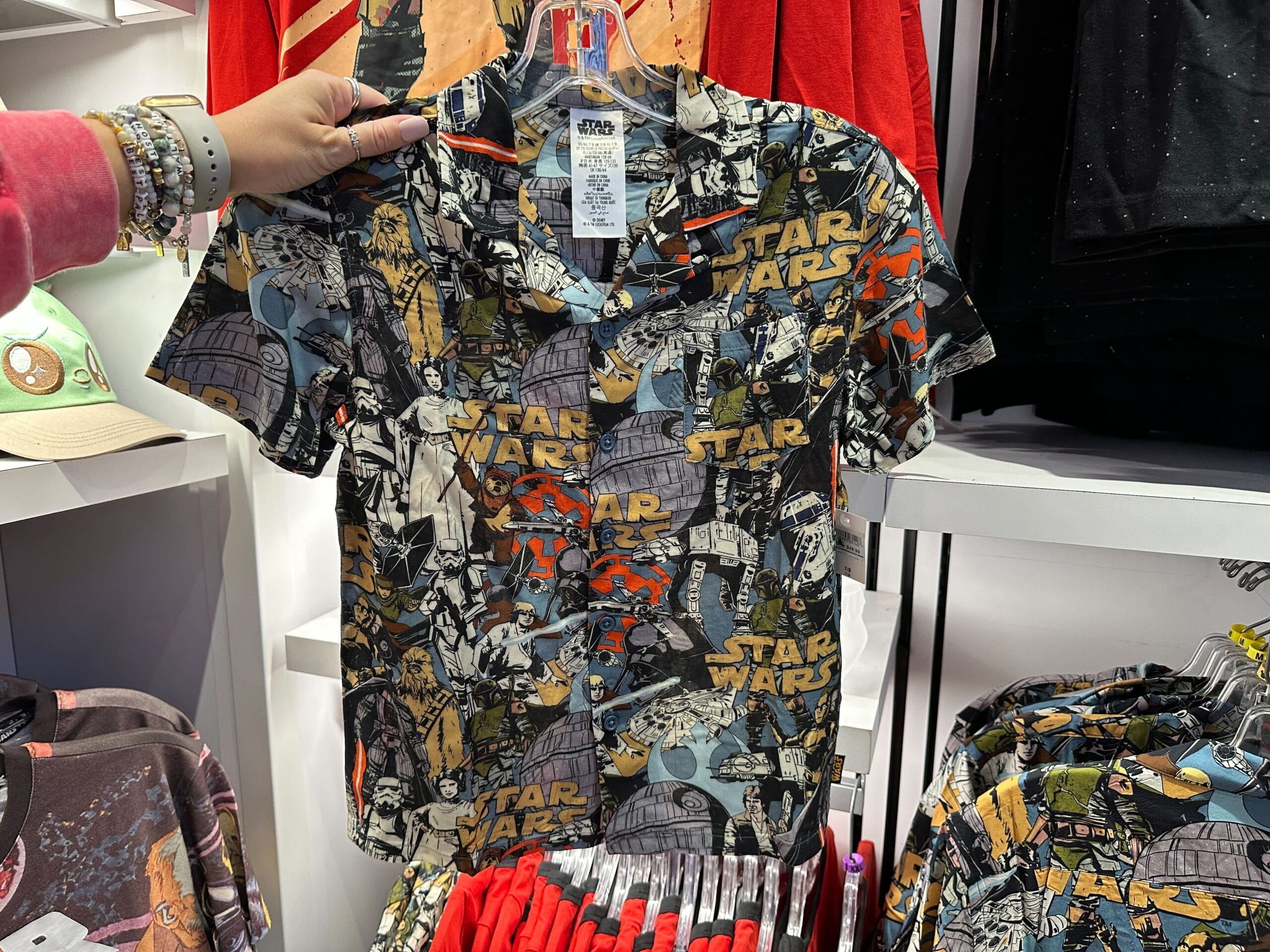 A hand holds a Star Wars shirt with comic-style graphics in a Disney Parks shop, surrounded by themed Disney merchandise displays.