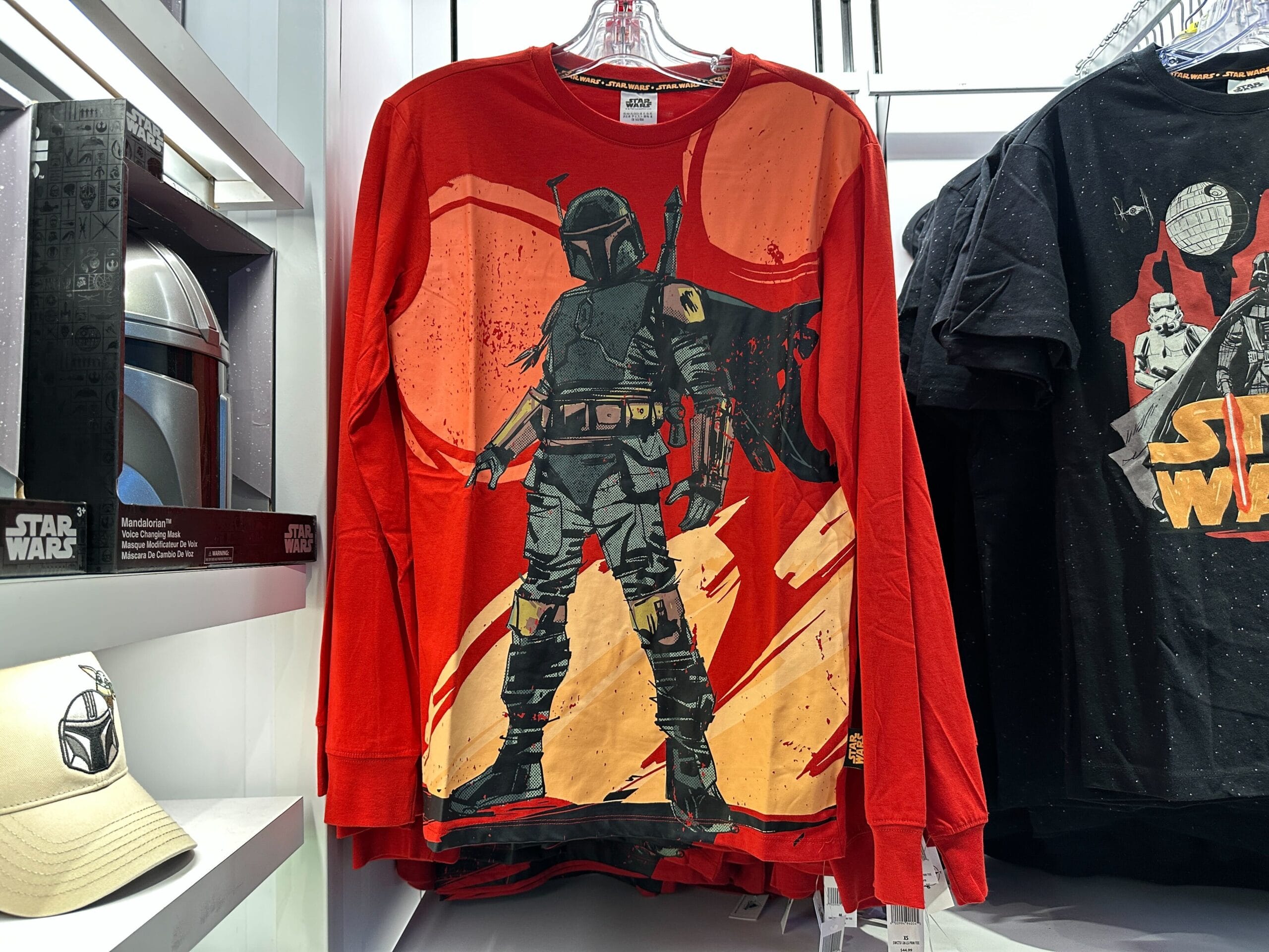 A red long-sleeve Boba Fett shirt hangs with Star Wars apparel on a store rack, likely found in a Disney park’s shop.