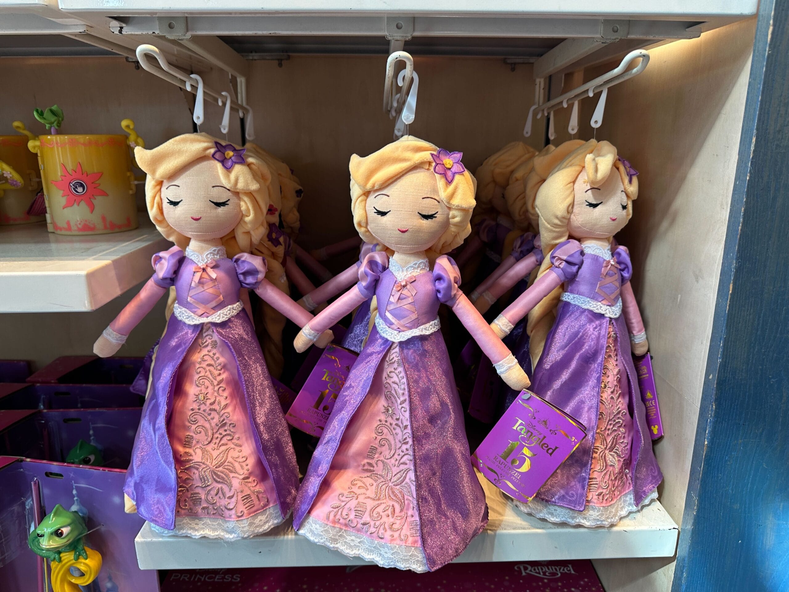 Three Rapunzel plush dolls in purple dresses hang on display in a Disney park store, evoking the Tangled castle’s whimsy.