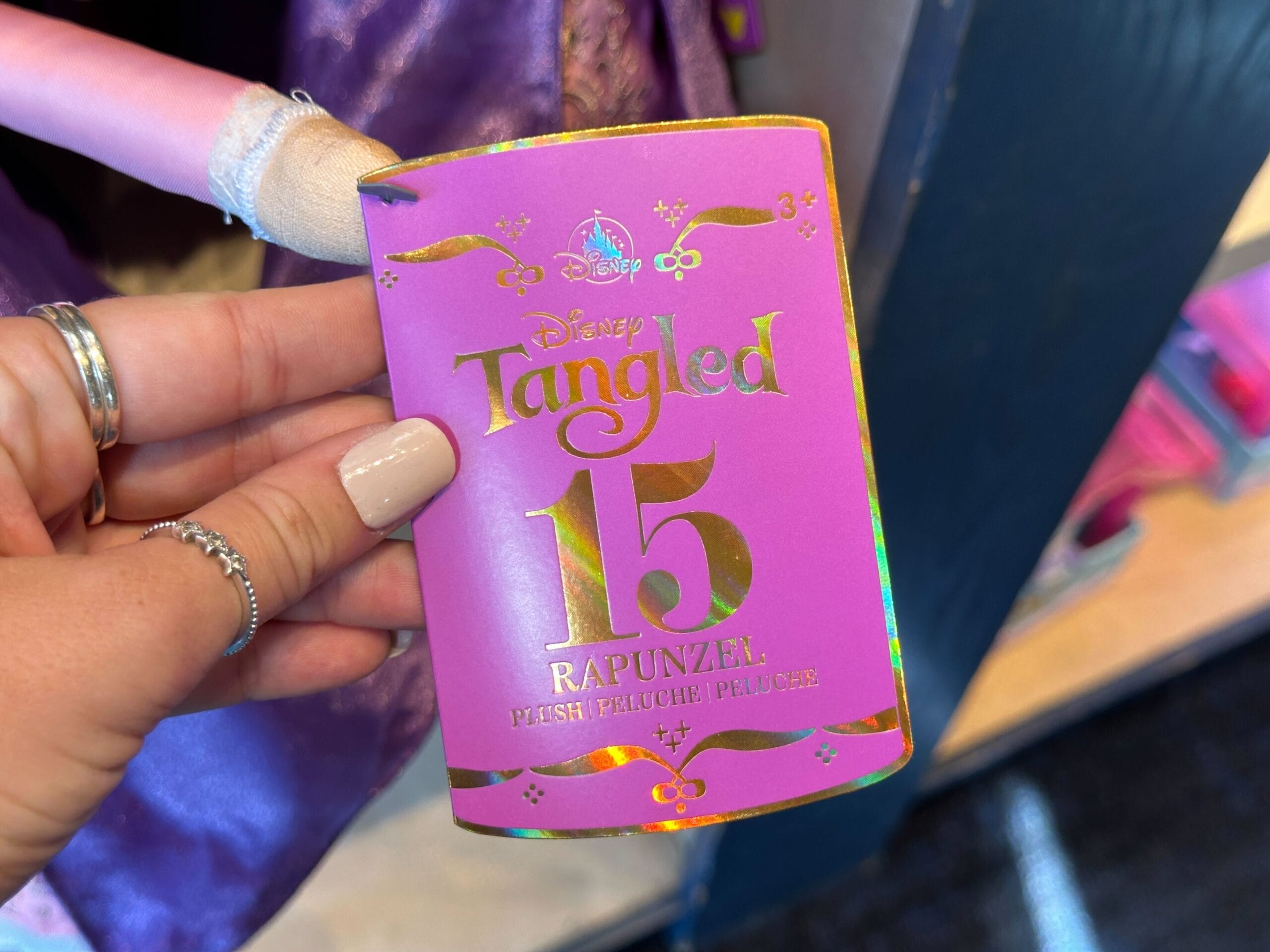 A hand holds a pink Disney Tangled tag reading "15 Rapunzel Plush Peluche Peluche," reminiscent of park-exclusive plush tags.
