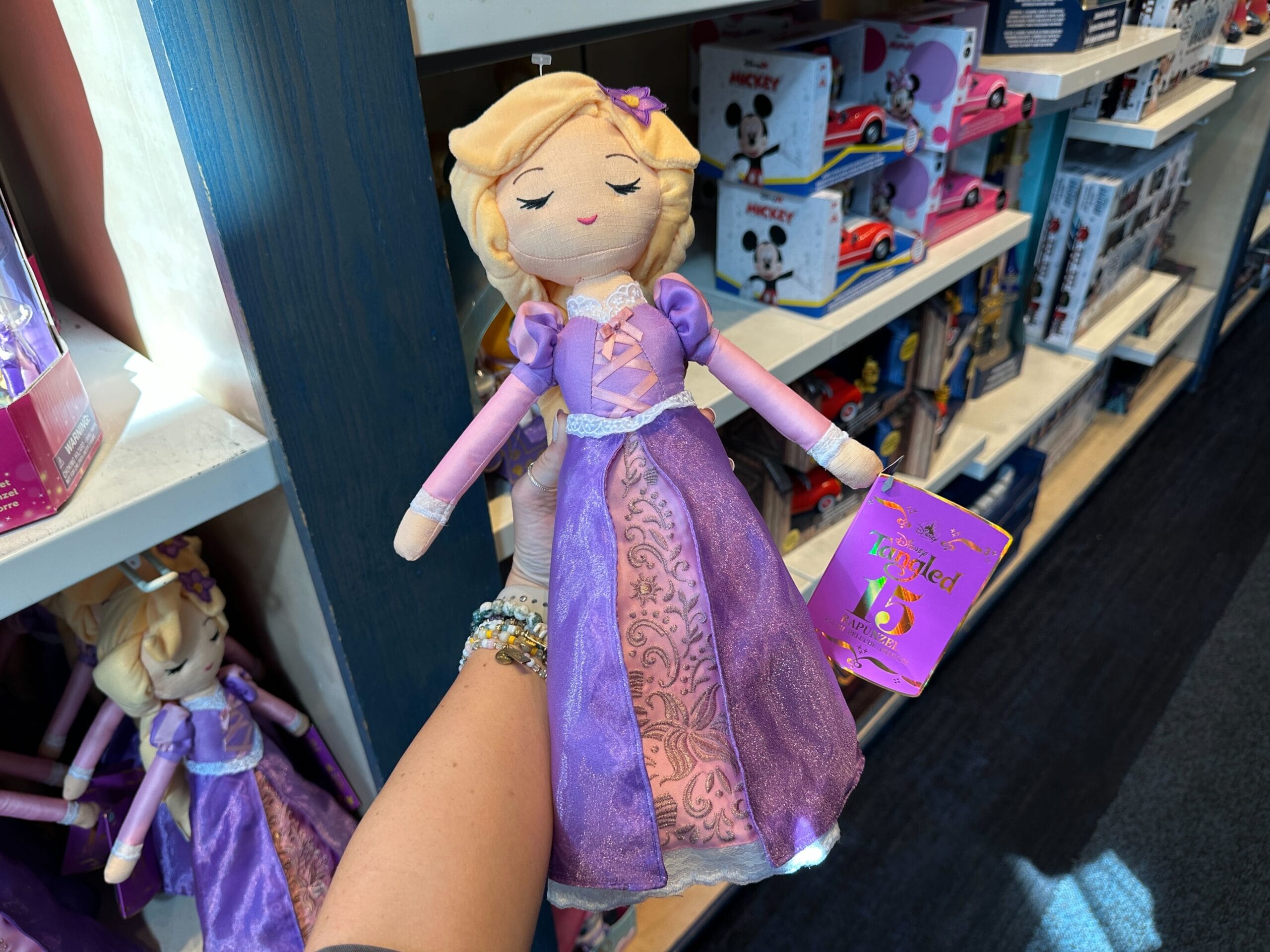 A hand holds a plush Rapunzel doll in a purple dress, showcased in a Disney park toy shop with themed shelves behind her.