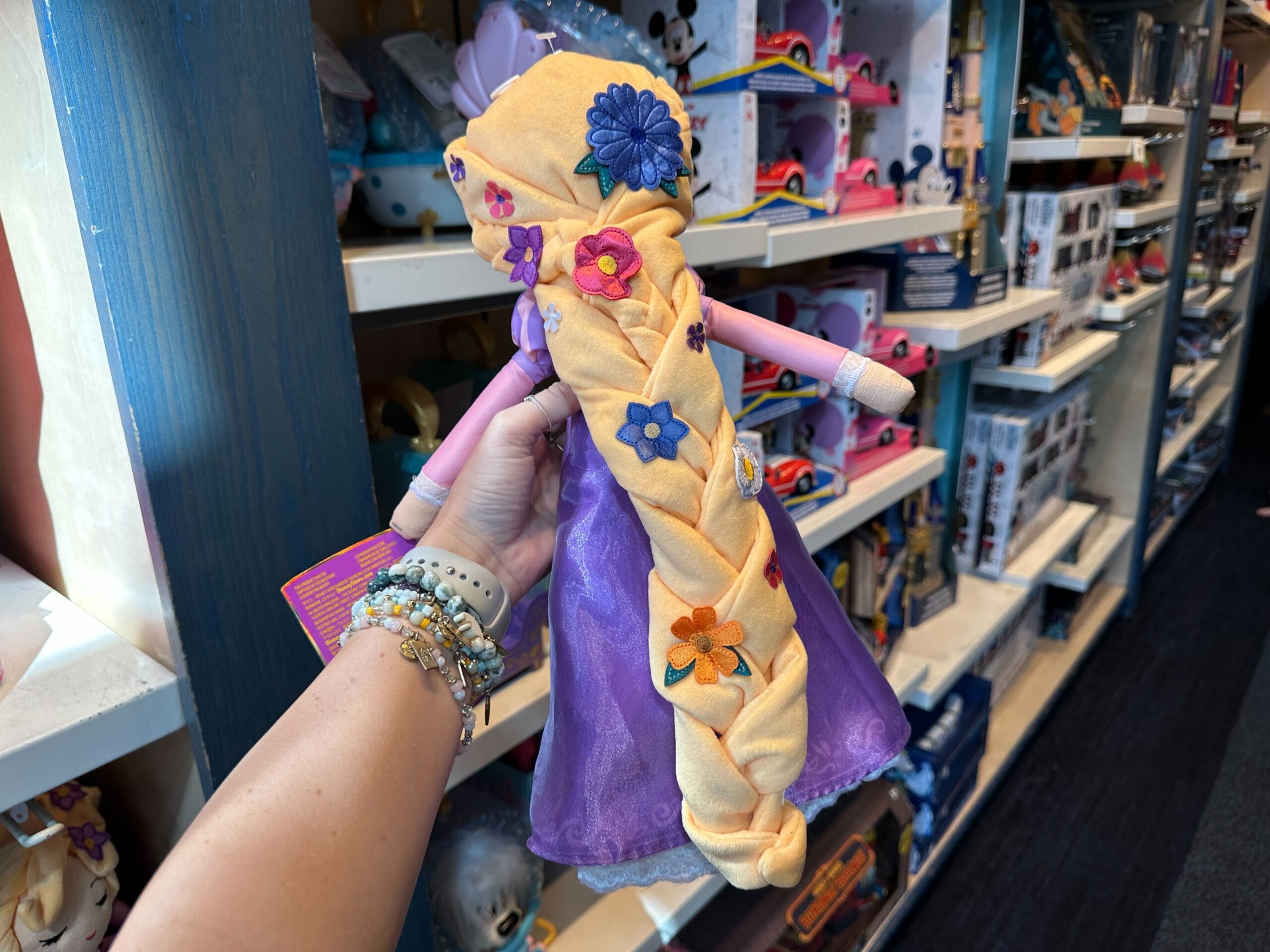 A person holds a Rapunzel doll with a long yellow braid and floral accents, browsing a Disney Park gift shop, not far from the castle.