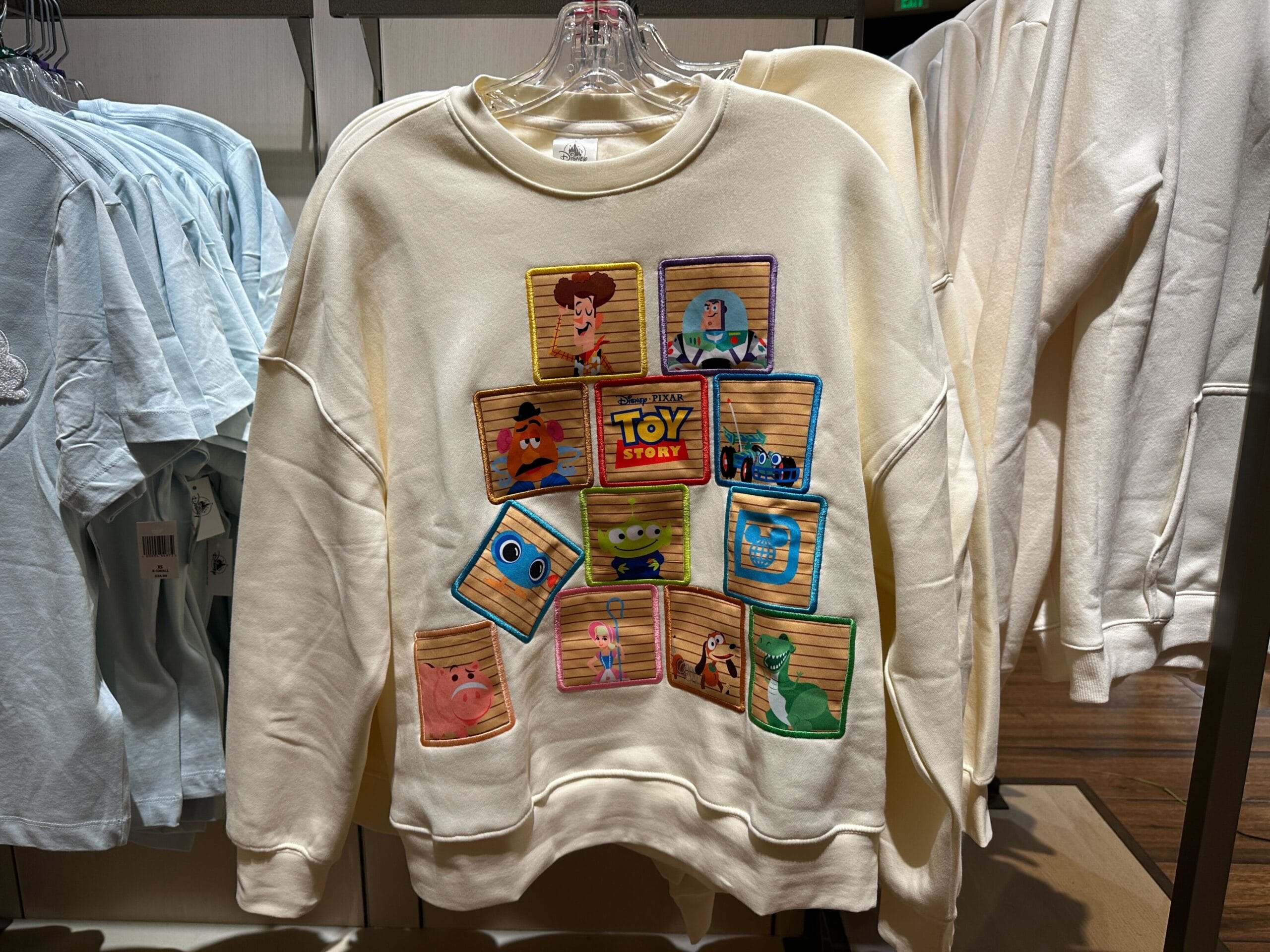 Cream Toy Story sweatshirt with colorful character portraits in box frames on the front, displayed in a Disney Park shop.